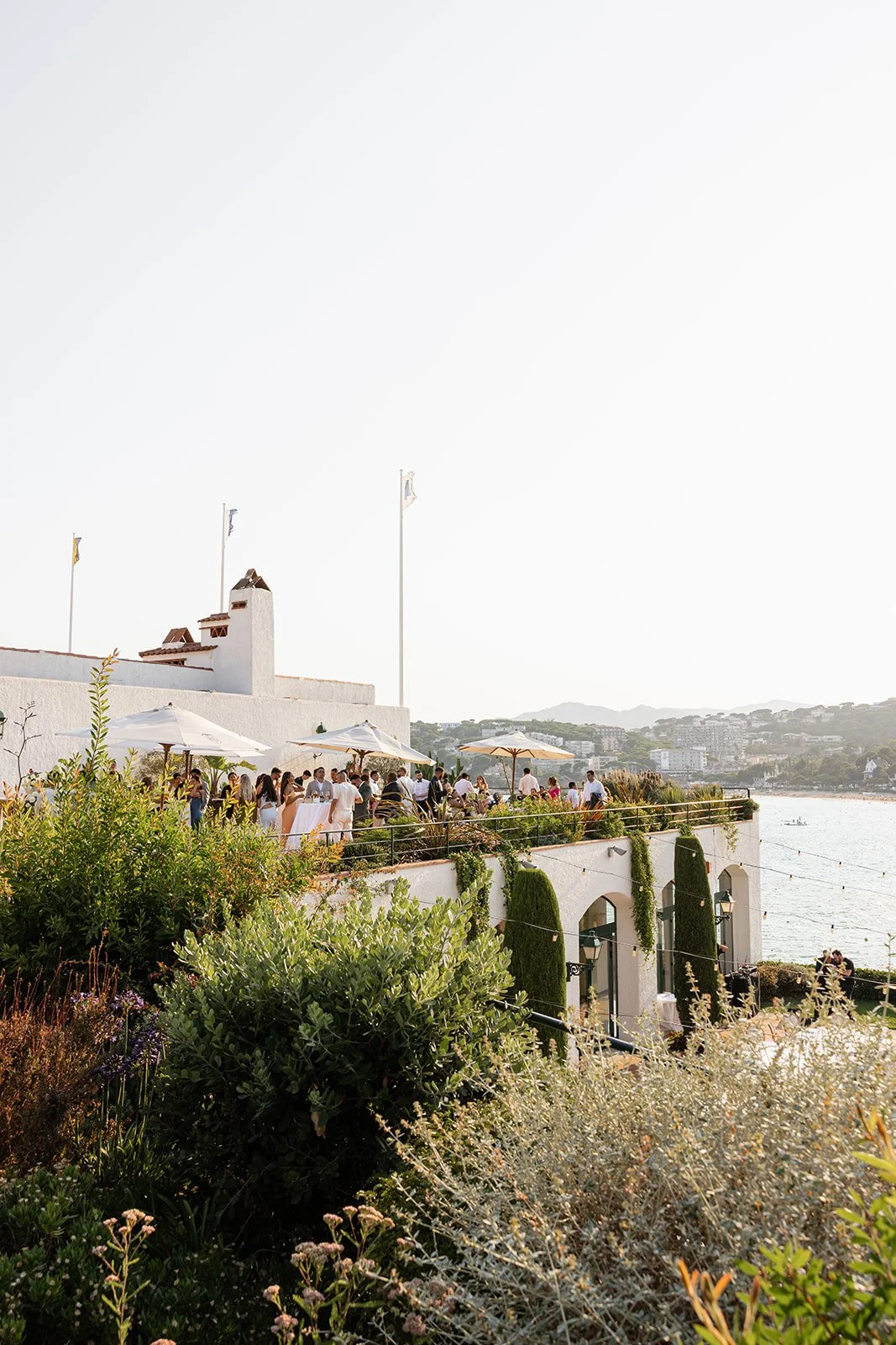 Outdoor gathering on a terrace by the water with umbrellas, guests, and scenic hills in the background.
