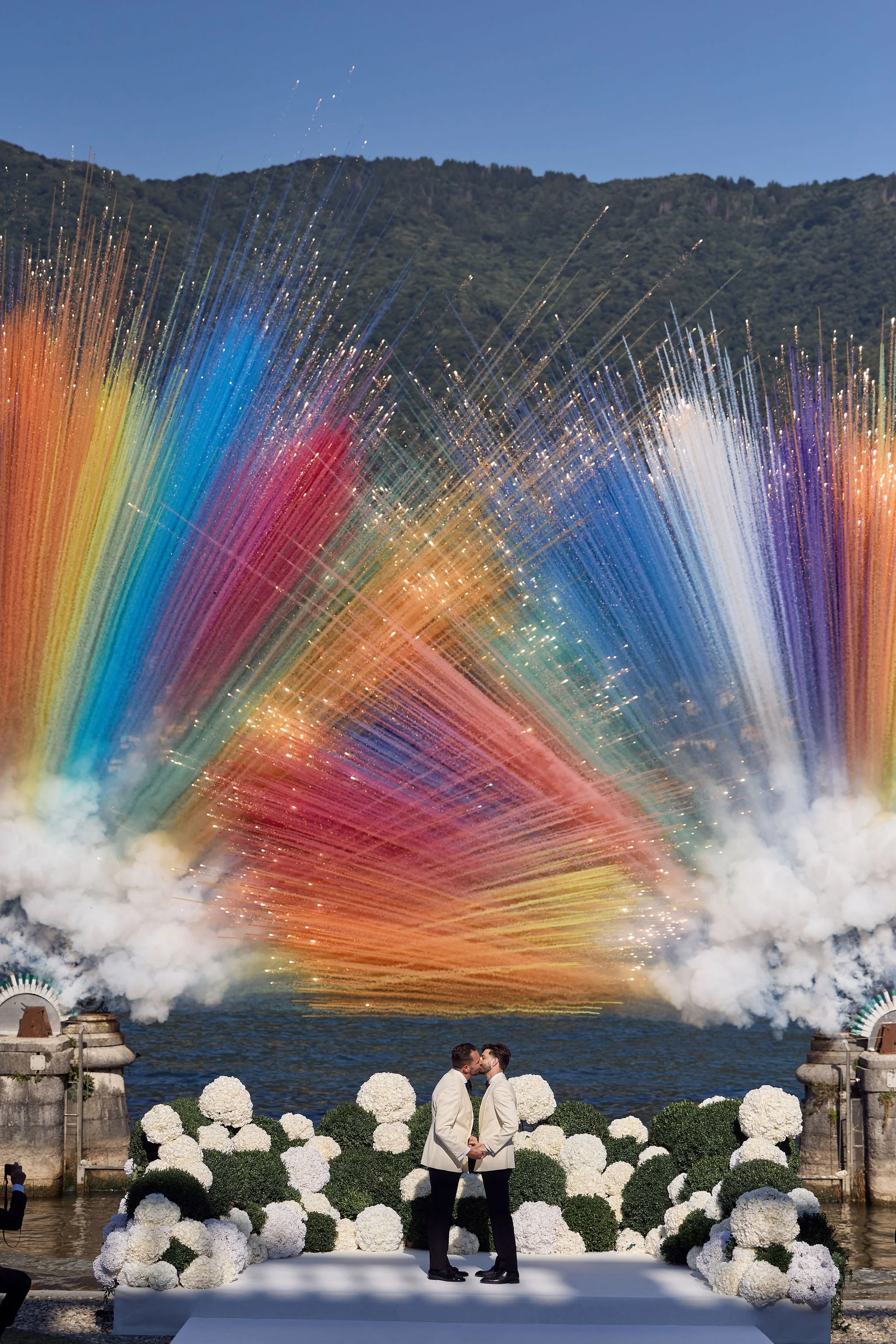 Couple in white tuxedos kissing beside the water beneath plumes of rainbow-colored smoke.