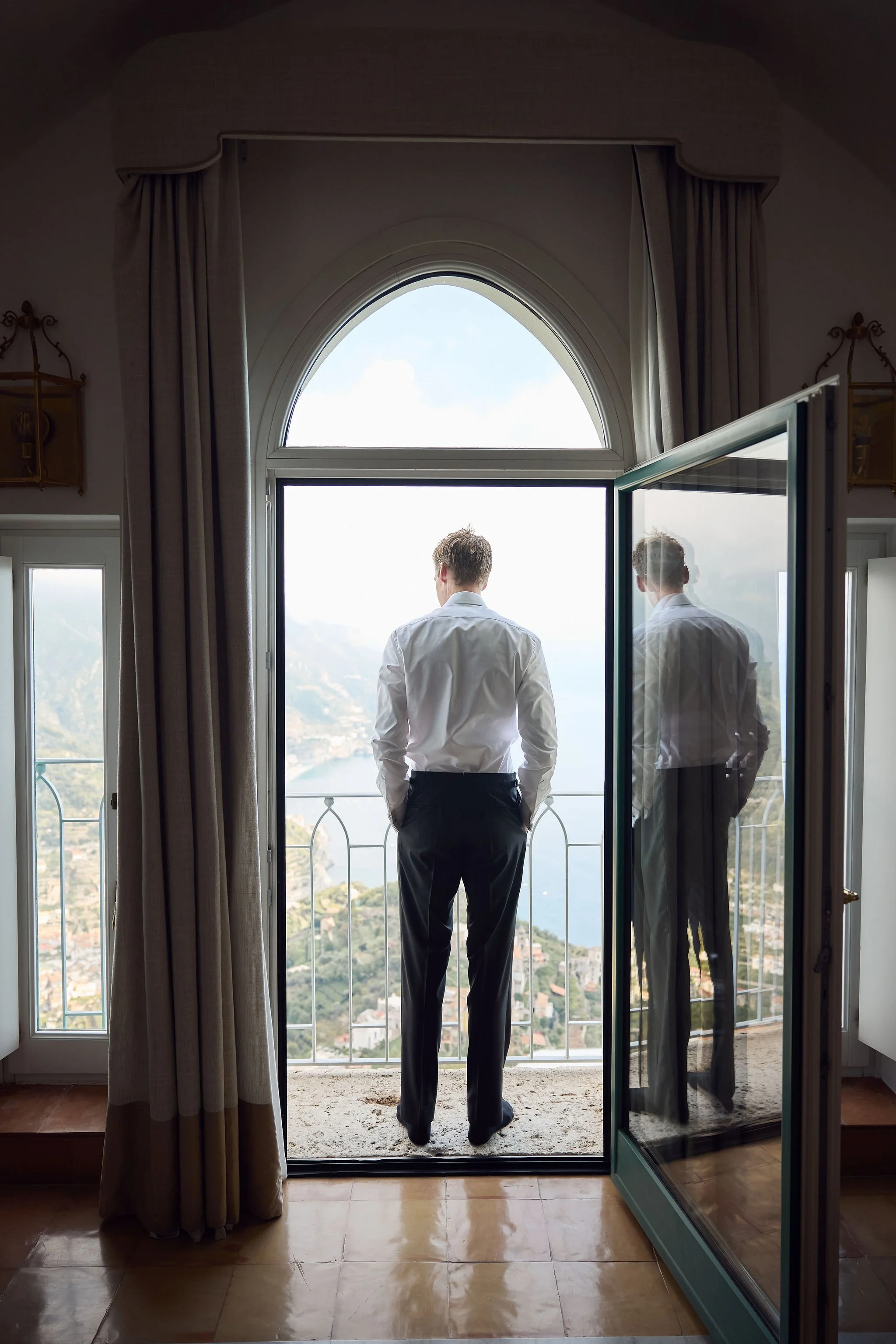 A man in a white shirt and black pants stands on a balcony, looking out at a scenic view of water and mountains. His reflection is visible in the glass door to the side.