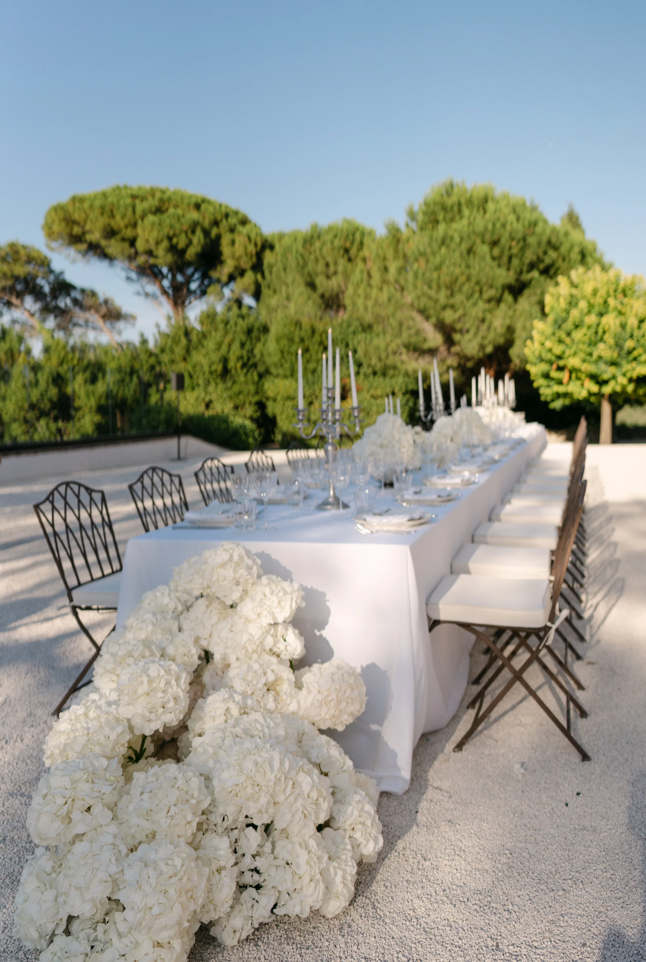 Outdoor wedding reception table with white tablecloth, floral centerpieces, and silver candelabras, surrounded by black chairs on a sandy surface, with tall green trees in the background under a clear blue sky.