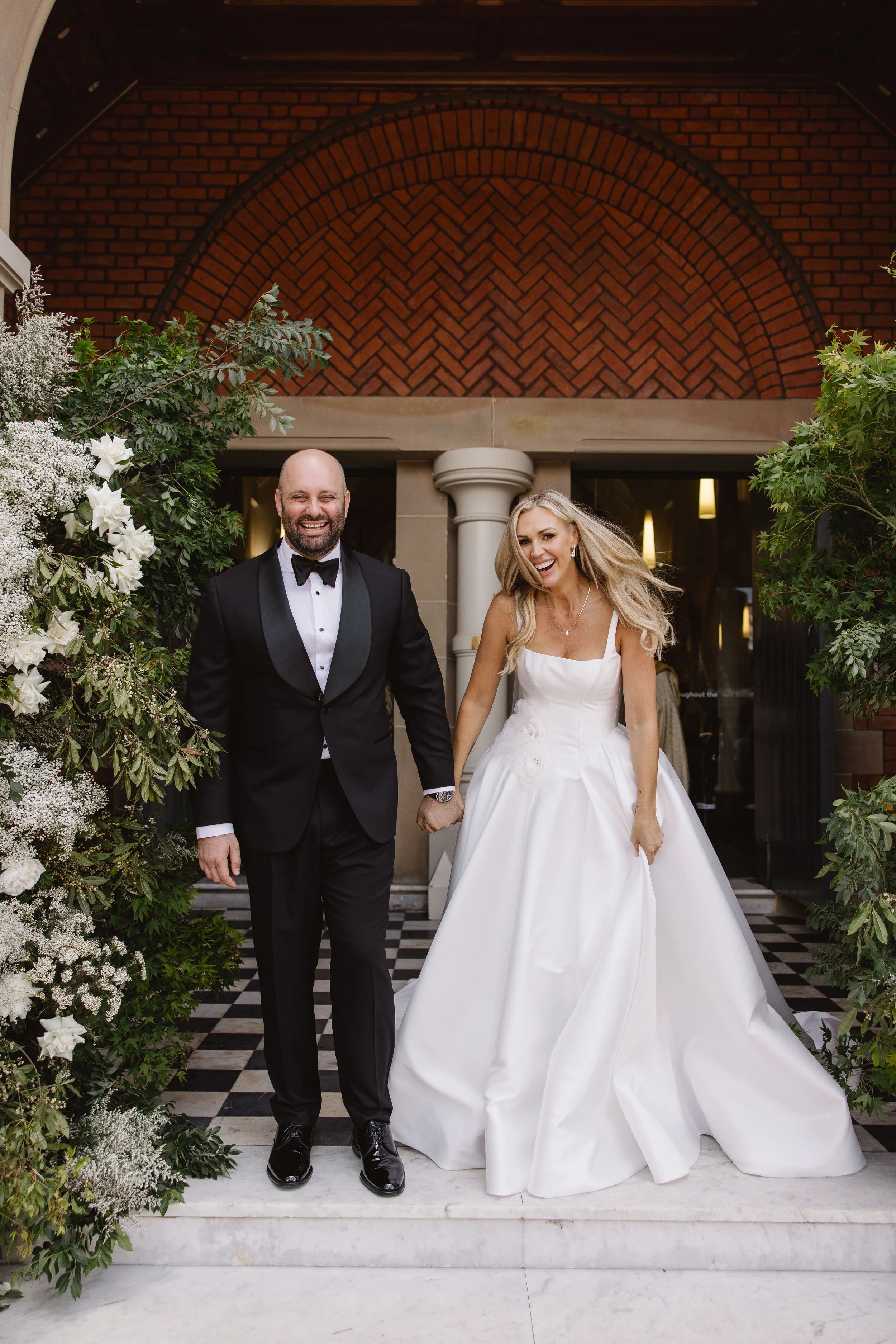A newlywed couple holding hands and smiling, standing outside on a black-and-white checkered floor, surrounded by lush green and white floral arrangements, with a brick and stone building in the background.