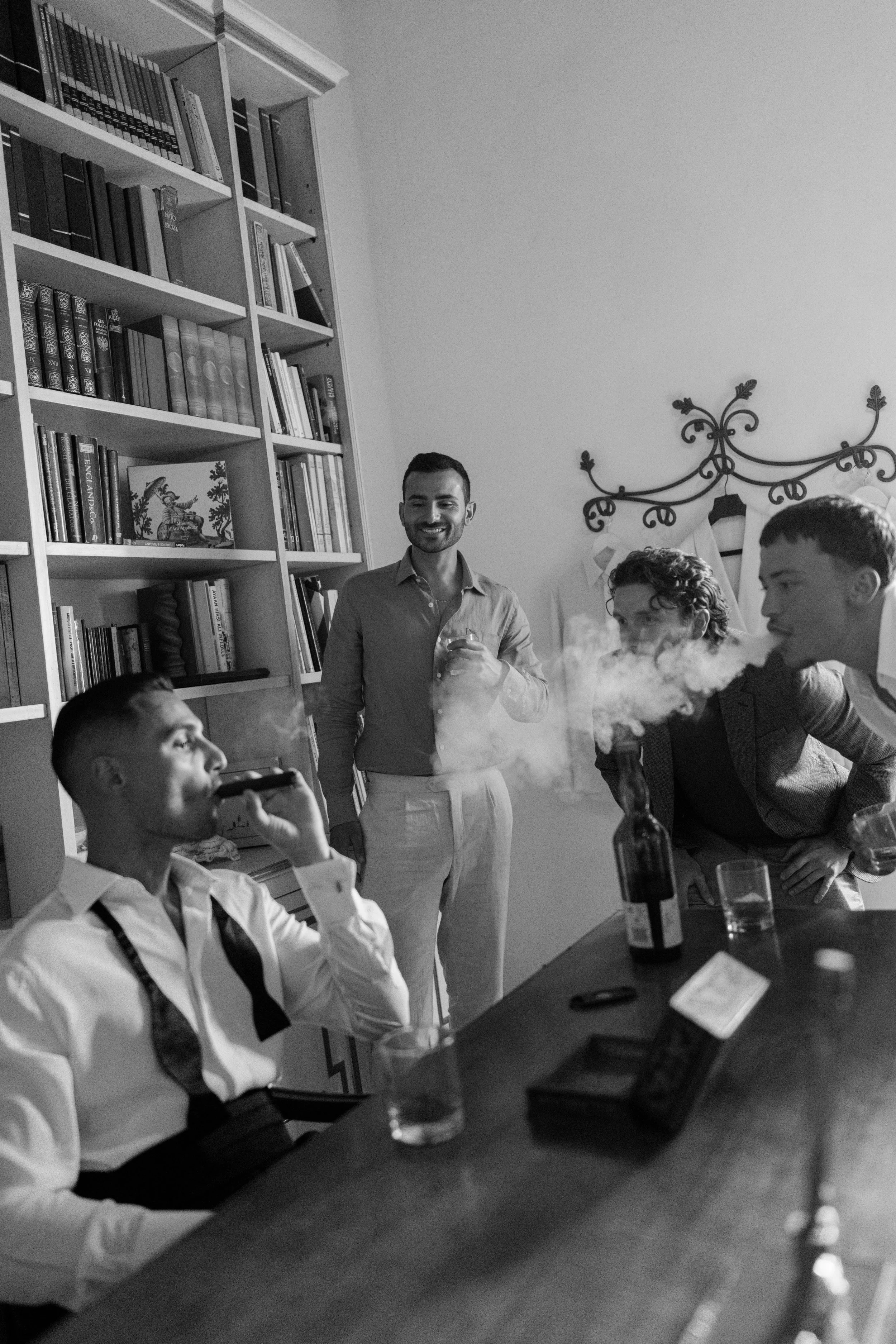 A group of young men with light to medium skin tones gathered around a table in a room with bookshelves. One man is standing and smiling while holding a drink. Two other men are seated, one drinking from a bottle and the other with curly hair, leanin