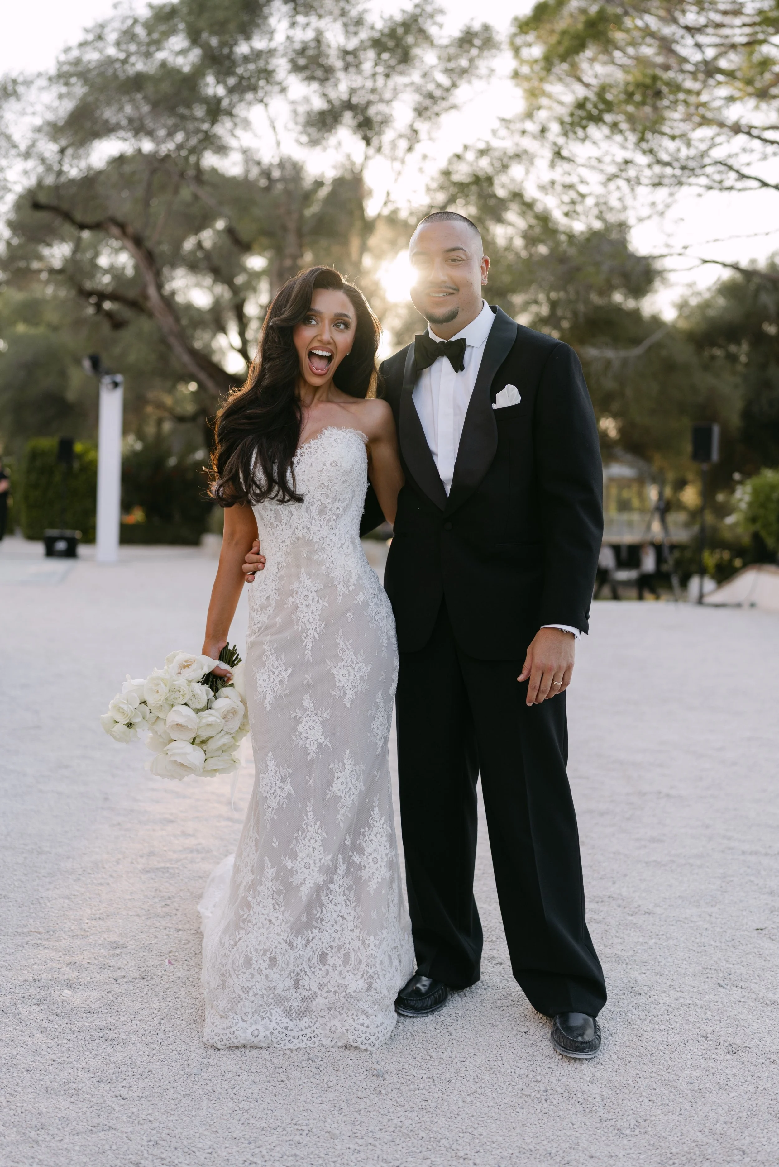 A bride and groom standing outdoors during sunset, with trees in the background. The bride is wearing a white, lace wedding dress and holding a bouquet of white roses. The groom is dressed in a black tuxedo with a bow tie. Both are smiling and appear