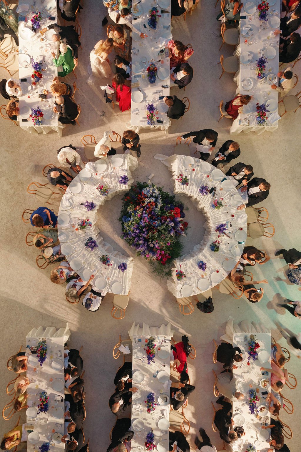 Overhead view of a banquet hall with round tables decorated with colorful flower arrangements, where guests are seated and mingling at a formal event.