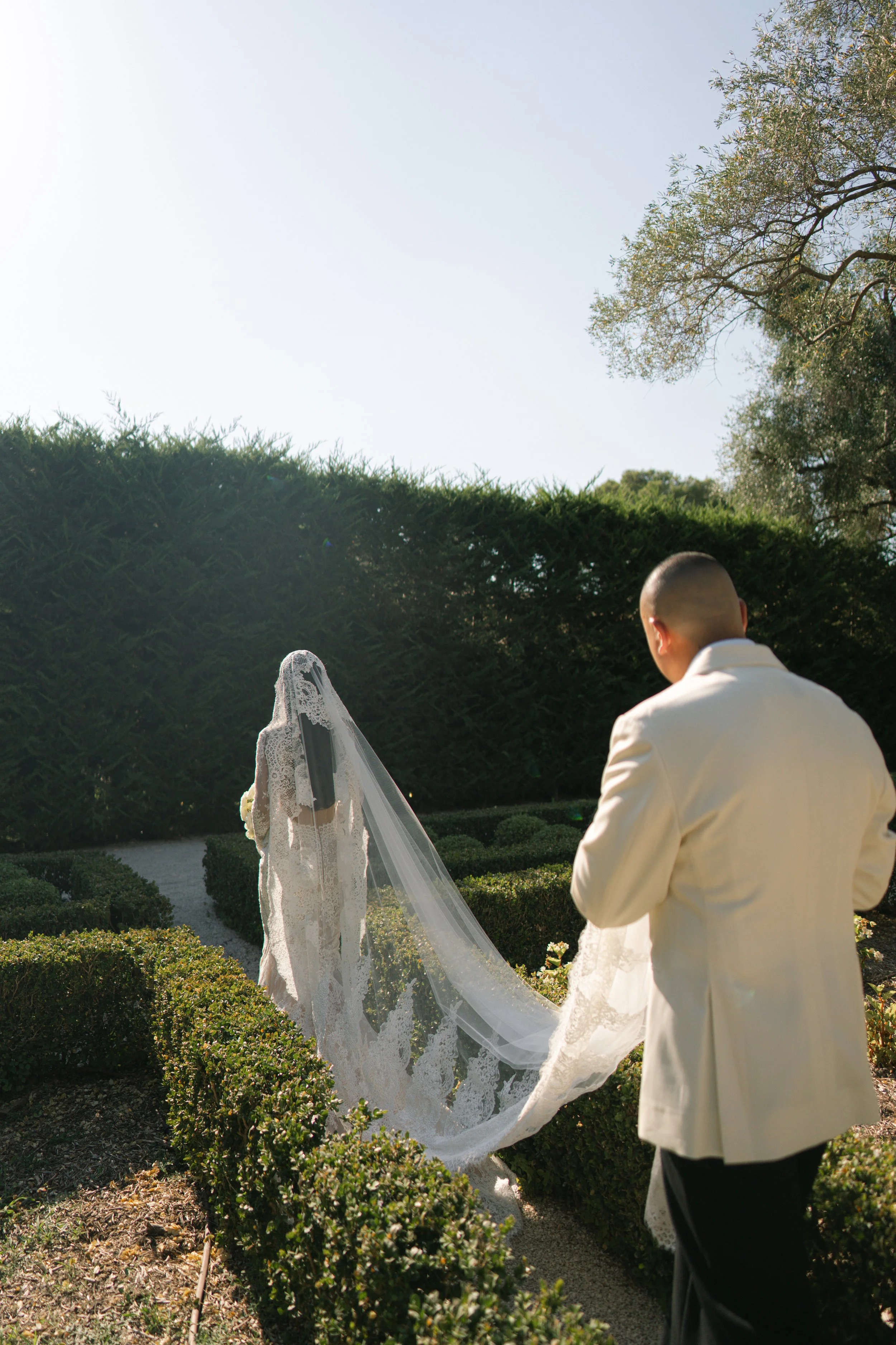 A bride and groom are standing outdoors in a garden with green bushes and trees, during a wedding ceremony. The bride is wearing a lace wedding dress with a veil, and the groom is dressed in a cream-colored suit. The groom is facing the bride, who is