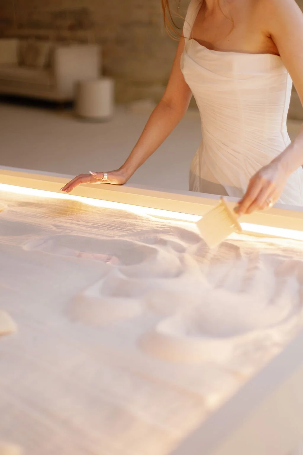 Person in a white strapless dress using a wooden spatula to smooth sand on a table.
