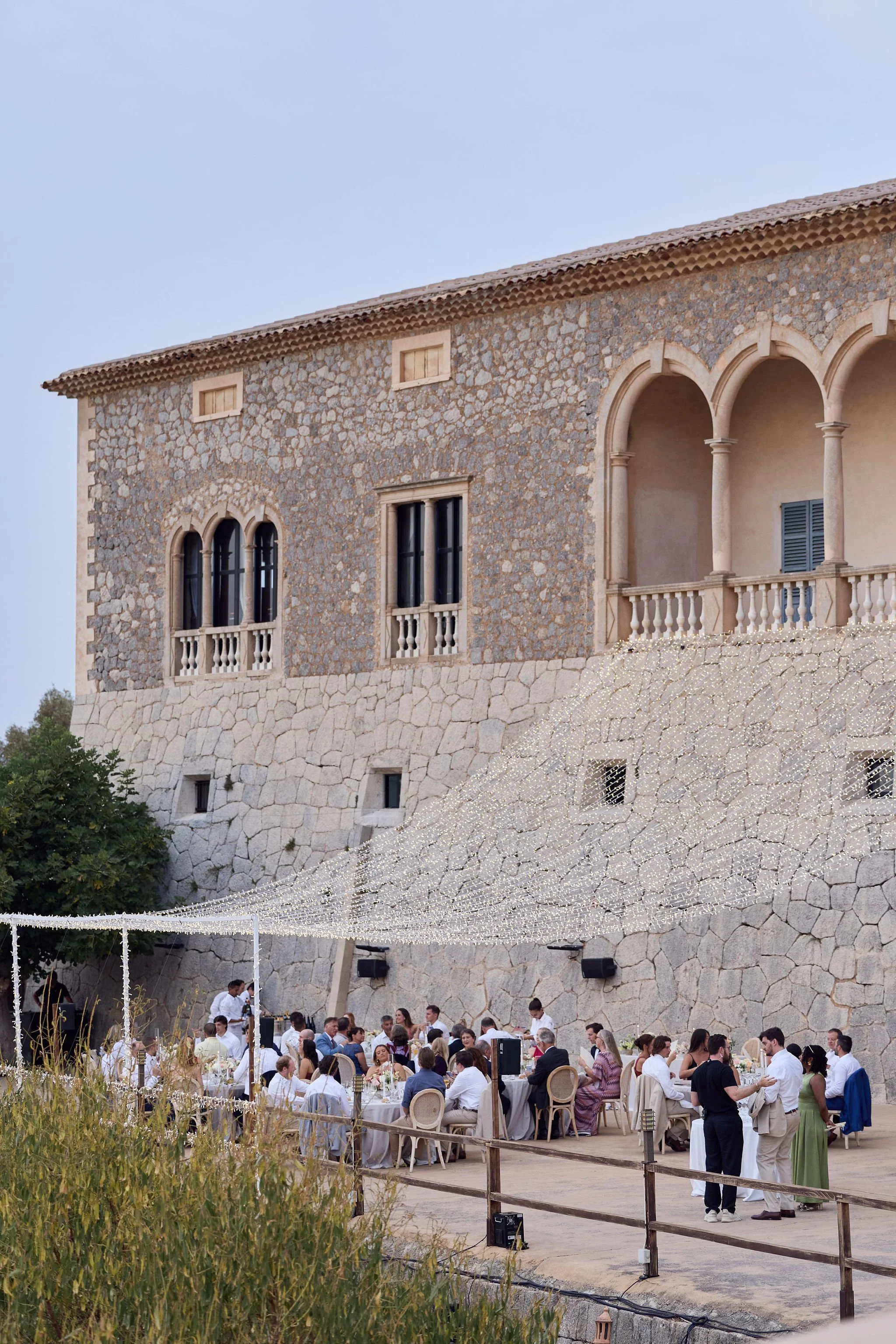 Wedding guests dining at round tables beneath string lights in a stone courtyard at dusk.