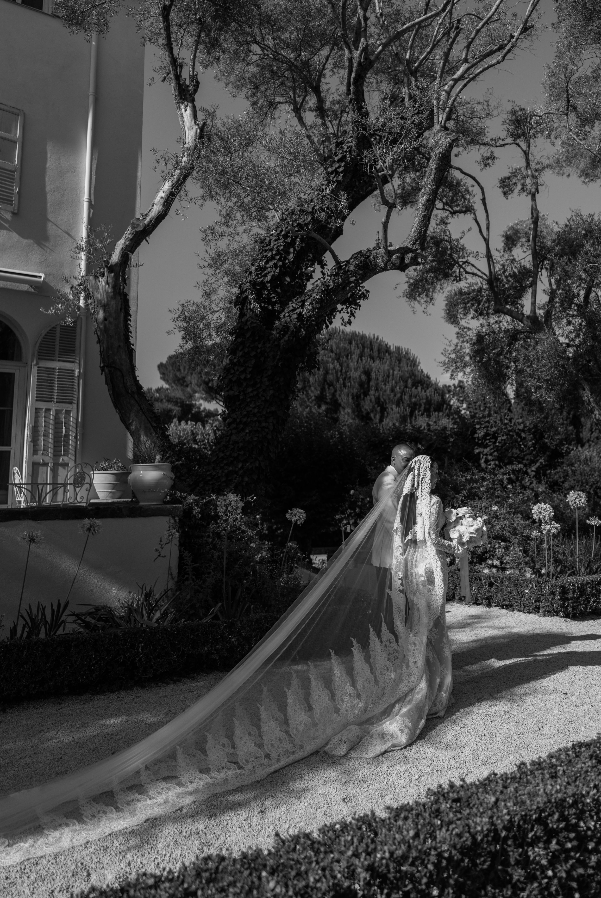 A bride and groom standing outdoors near a large tree, with the bride wearing a long lace wedding dress and veil, and the groom dressed in a suit, during a wedding ceremony.