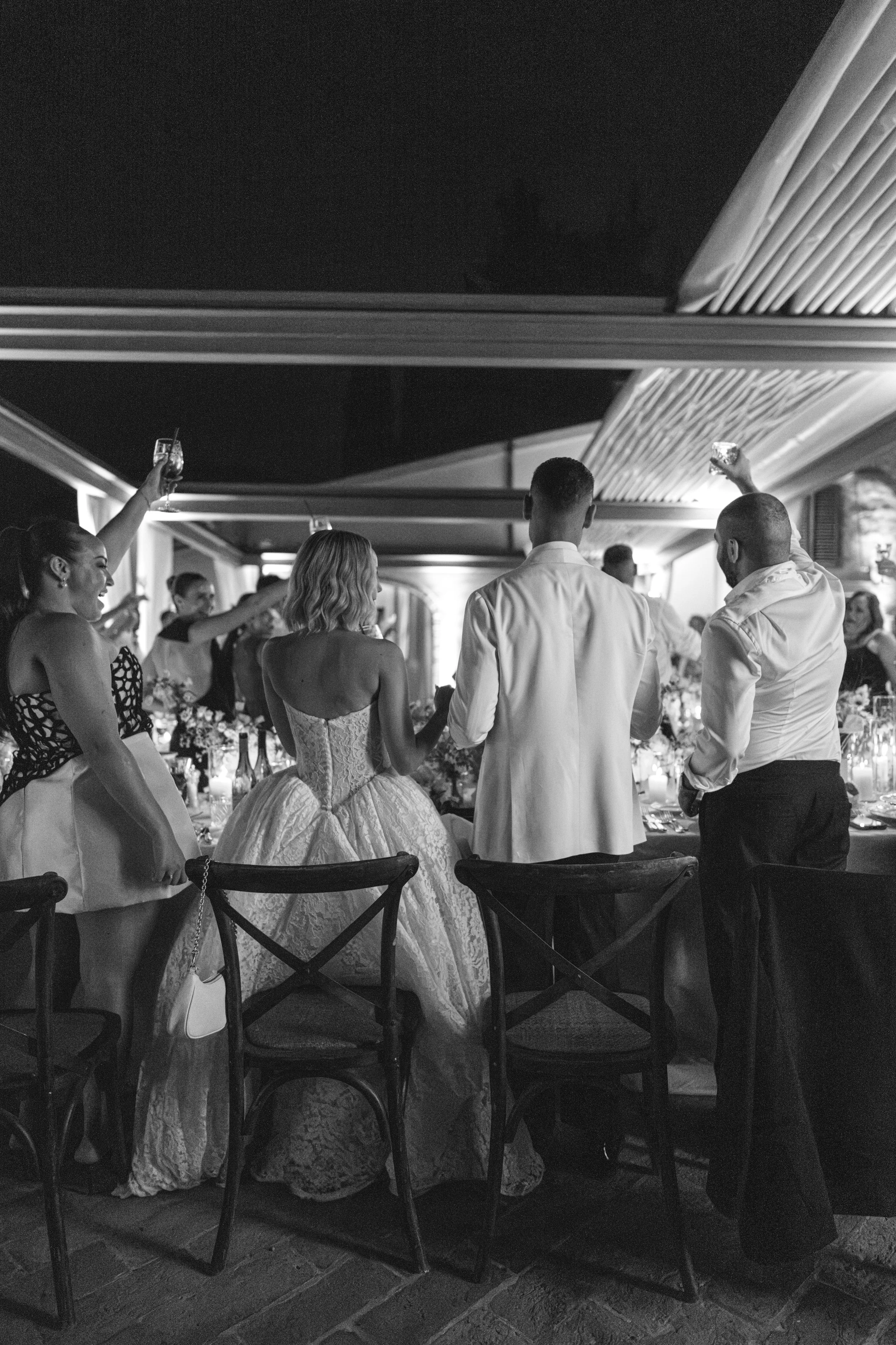 Black and white photo of guests at a wedding reception raising glasses in a toast, with the bride and groom in the center.