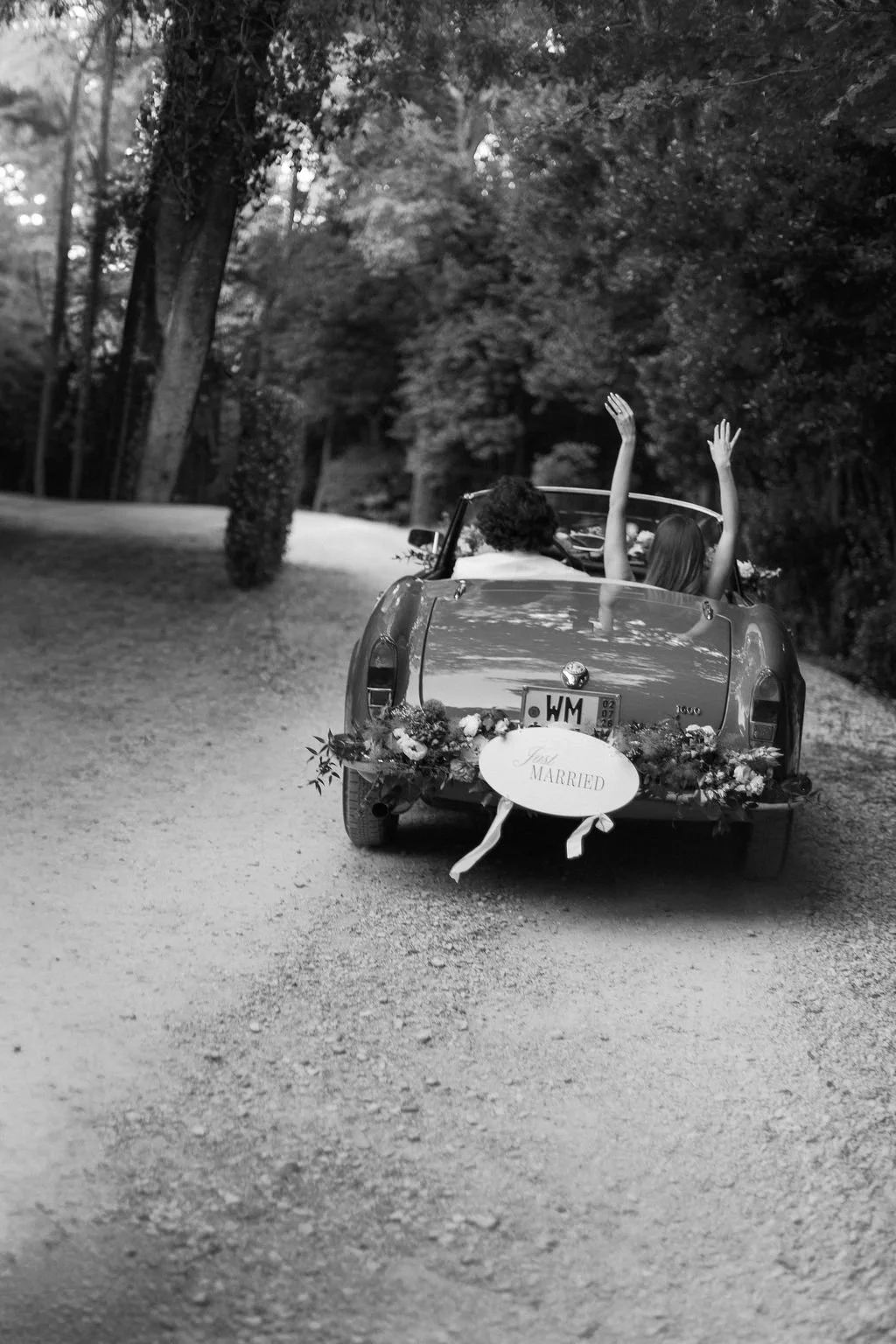A black and white photo of a vintage convertible car decorated with flowers and a "Just Married" sign on the back. Two women are sitting inside the car with their arms raised in celebration. The car is on a dirt road surrounded by trees.