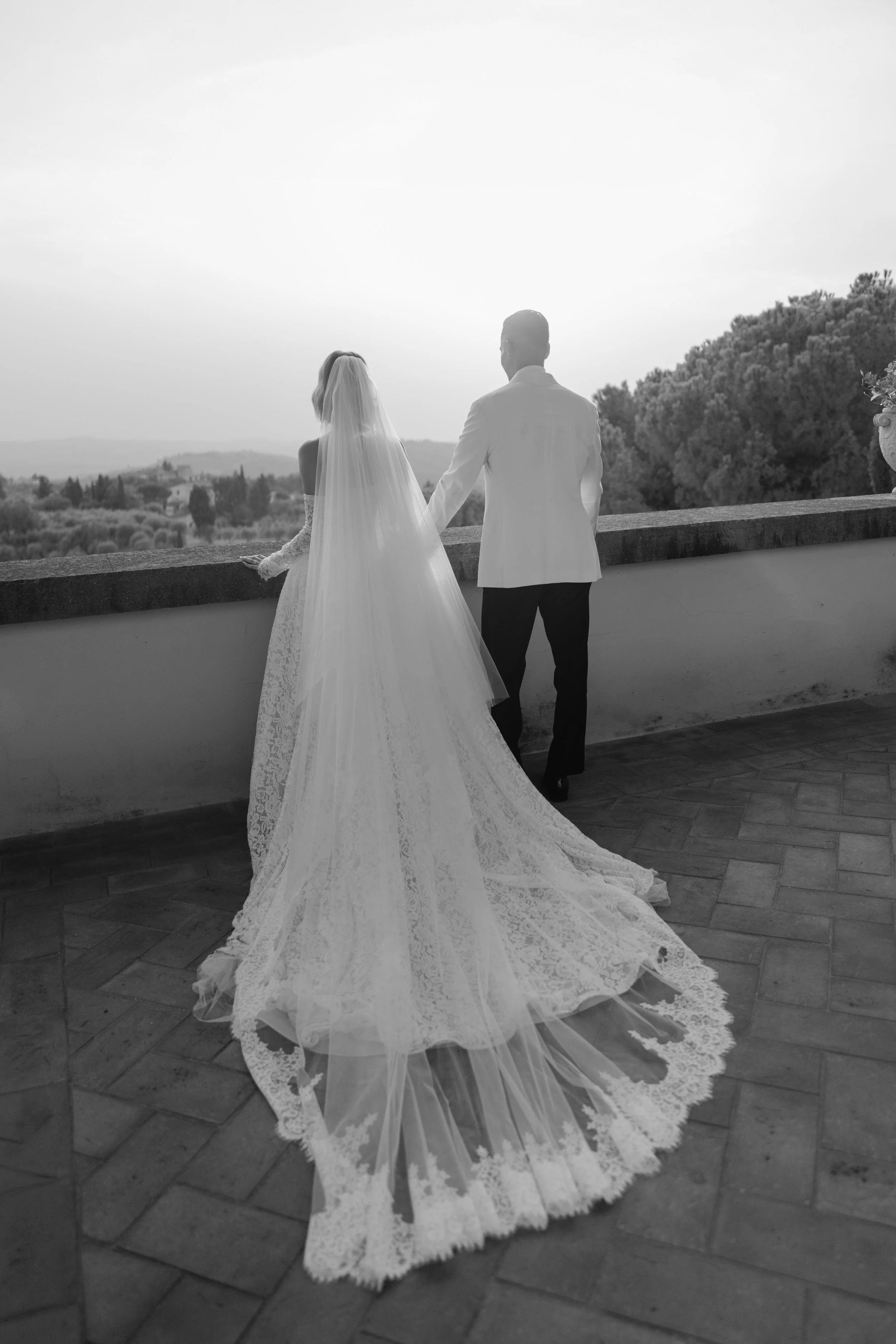 A bride and groom stand together on a terrace at sunset, the bride in a long lace wedding dress with a veil, and the groom in a white jacket and dark pants, overlooking a scenic landscape with trees and hills.
