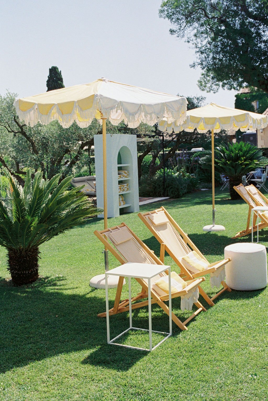 Outdoor lounge area with yellow striped umbrellas, wooden lounge chairs with pillows, a small white side table, lush green grass, and tropical plants.