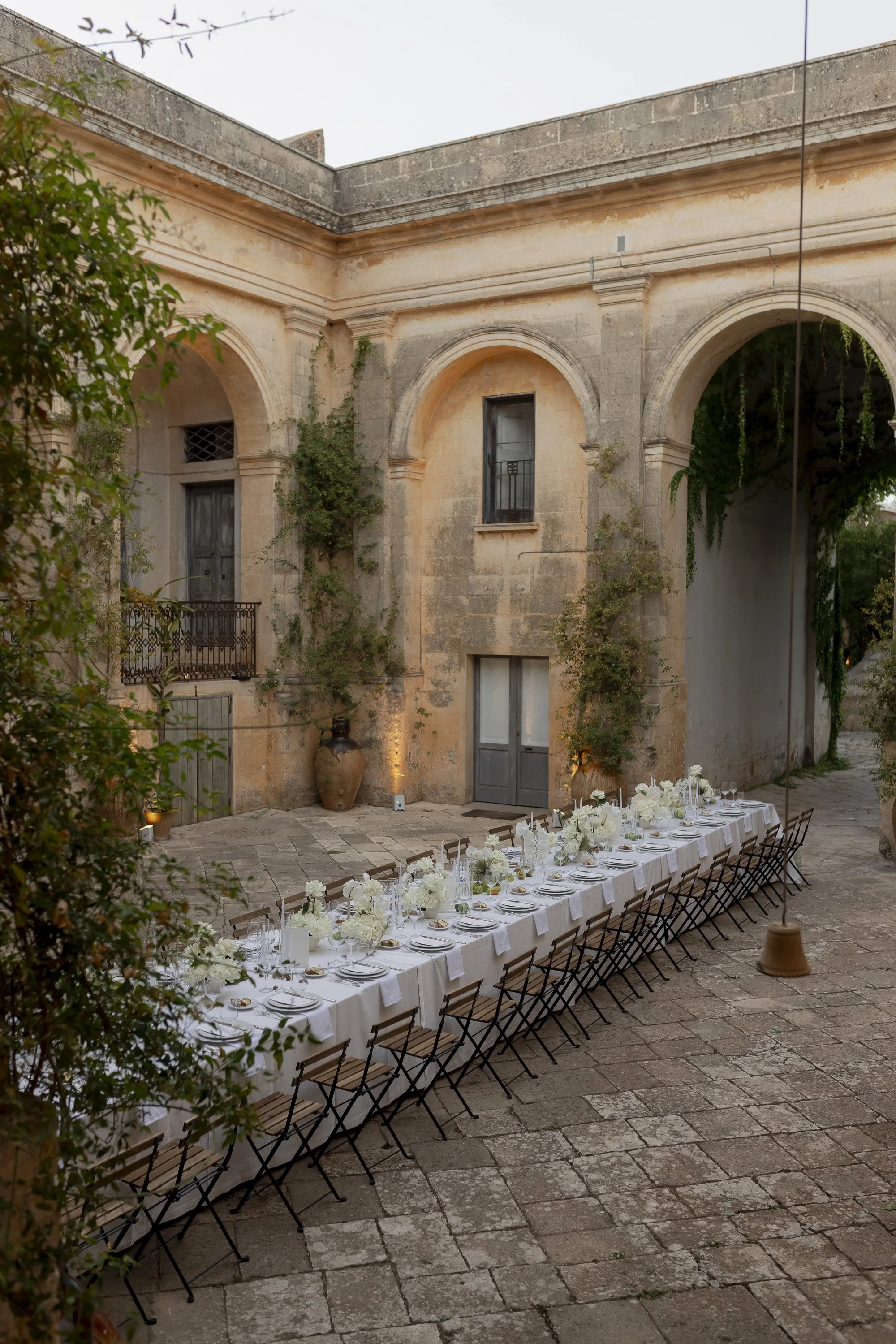 Long dining table with white linen, flowers and candles in a courtyard framed by stone arches and greenery.