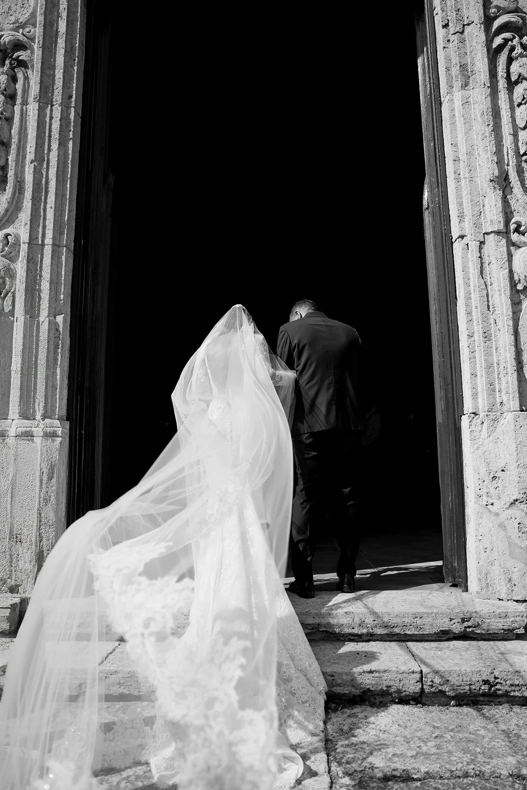 Bride in a wedding gown and veil walking into a church with a groom in a suit from behind.