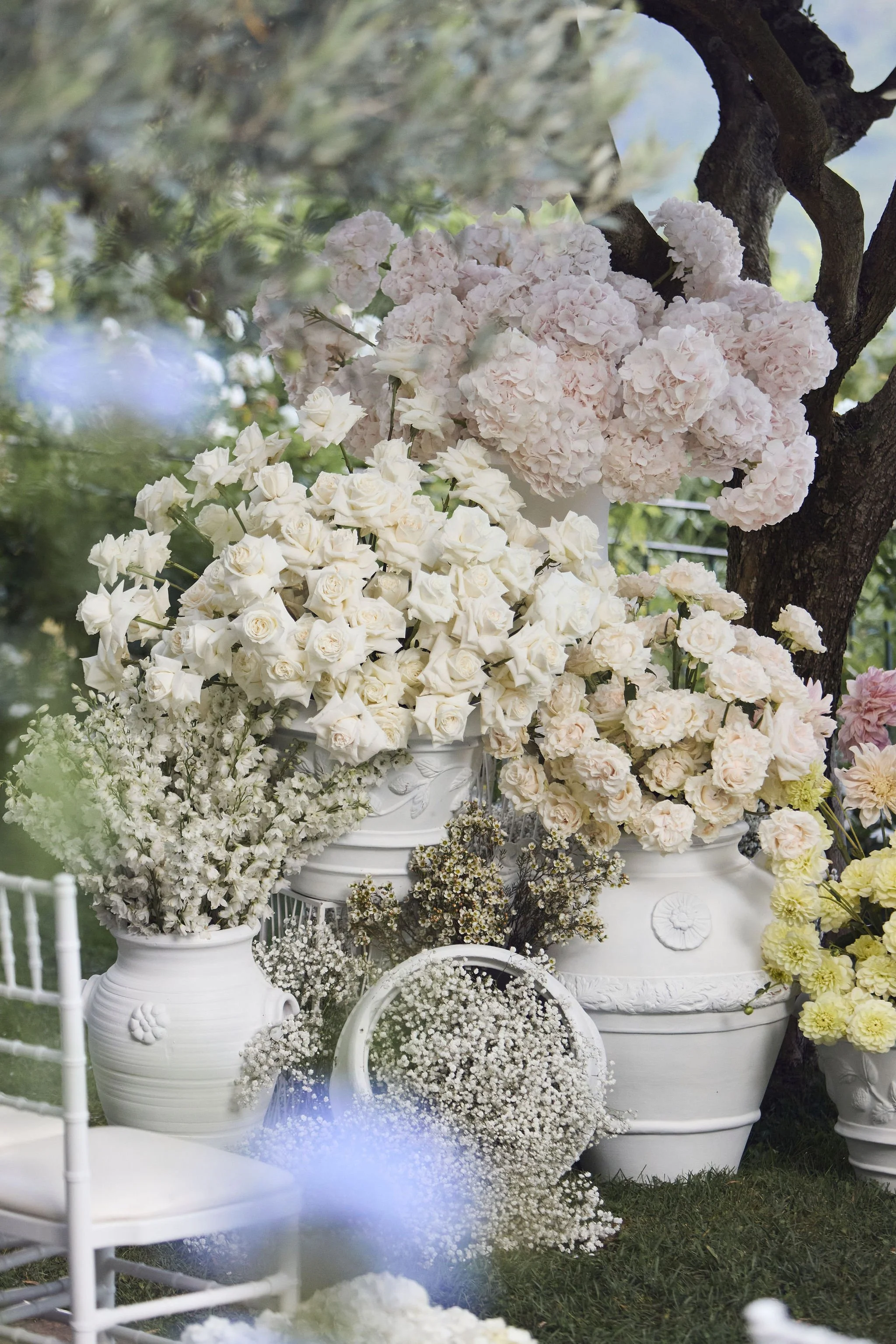 Flower arrangements with white and pink flowers placed in white ceramic vases and pots outdoors next to a tree.