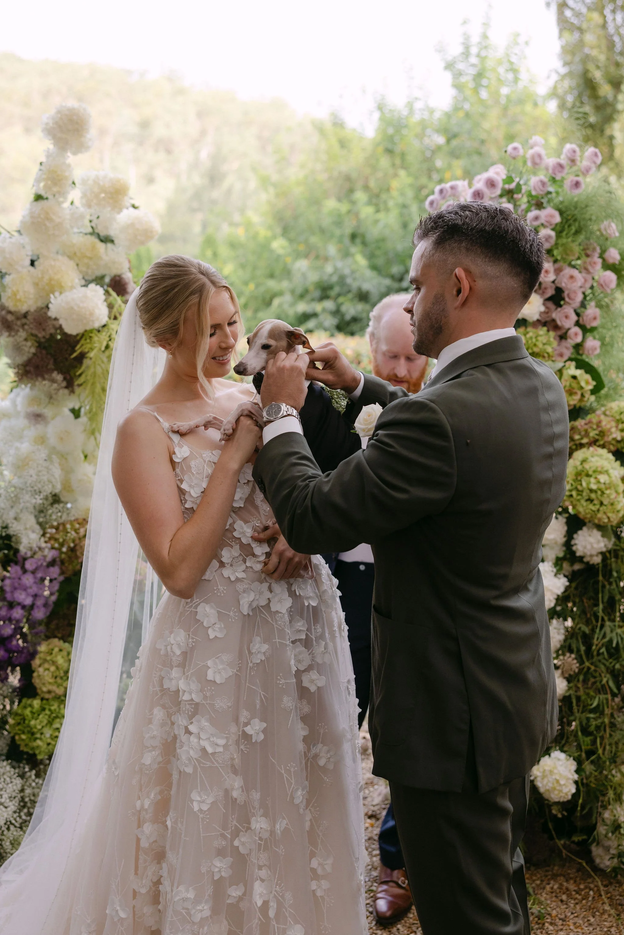 Bride and groom exchanging vows with a dog at their wedding ceremony outdoors, surrounded by colorful flowers and greenery.