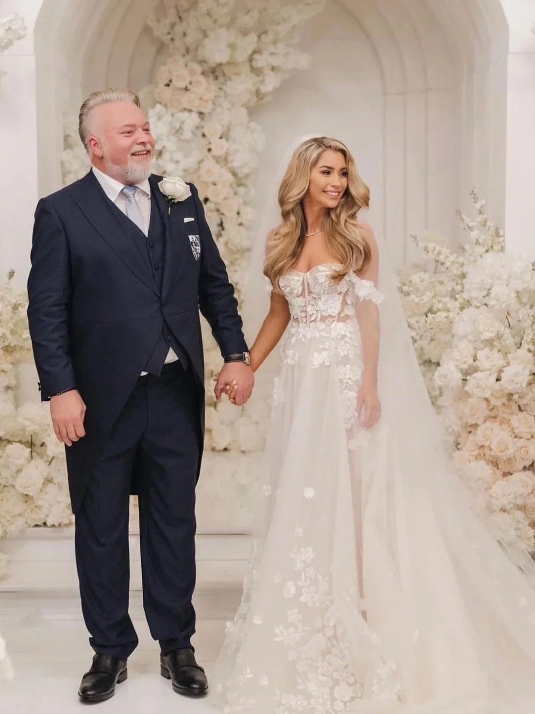 A bride and groom holding hands and smiling at their wedding ceremony, with a floral backdrop of white flowers.