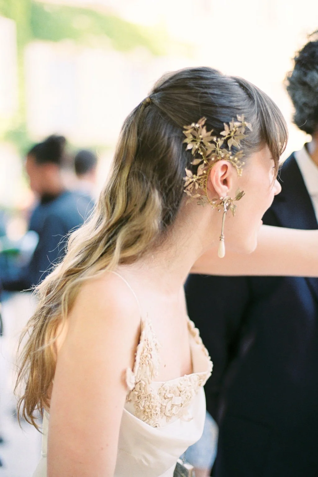 A woman with long, wavy hair wearing an elegant dress with floral embroidery, accessorized with gold leaf earrings and a gold hairpiece with leaves and a hanging pearl, at a social event.