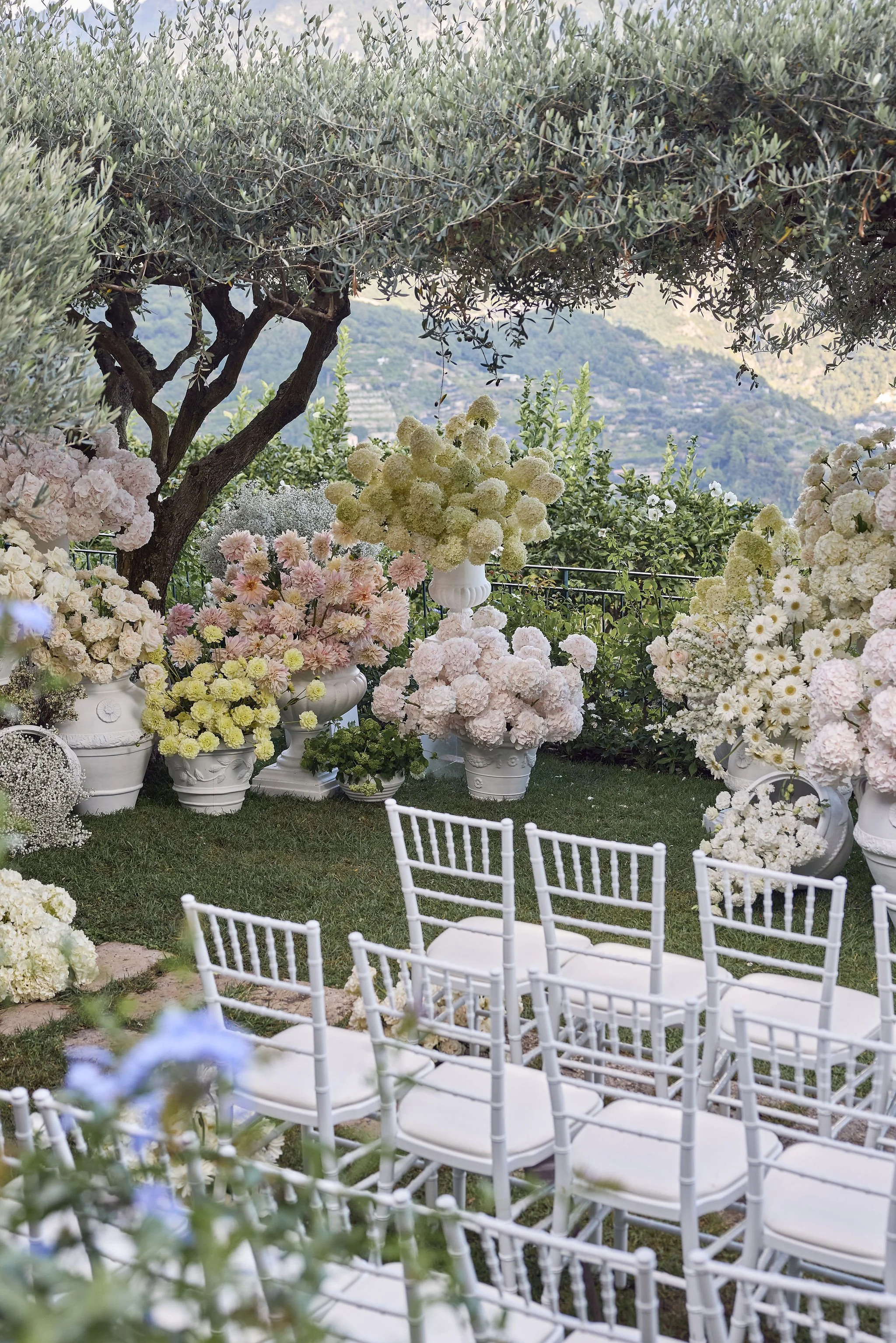 A beautifully decorated outdoor event space with white chairs arranged in rows, large white flower arrangements in decorative pots, and lush greenery including a large tree with a backdrop of distant hills.