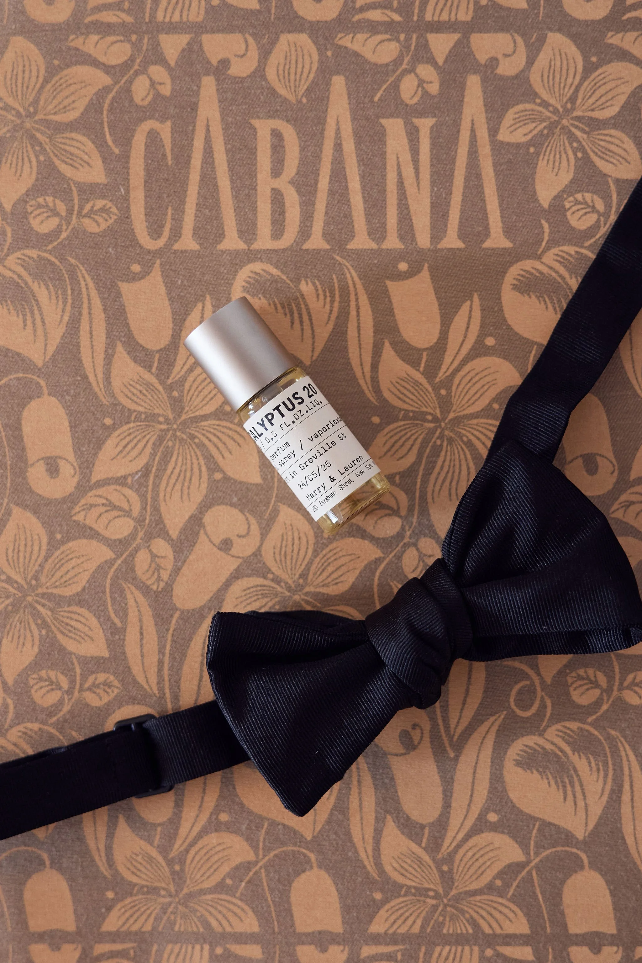 A small bottle of perfume labeled "HYPTUS 20," a black satin bow tie, and a decorative brown paper with a floral pattern and the word "CABANA" printed on it.