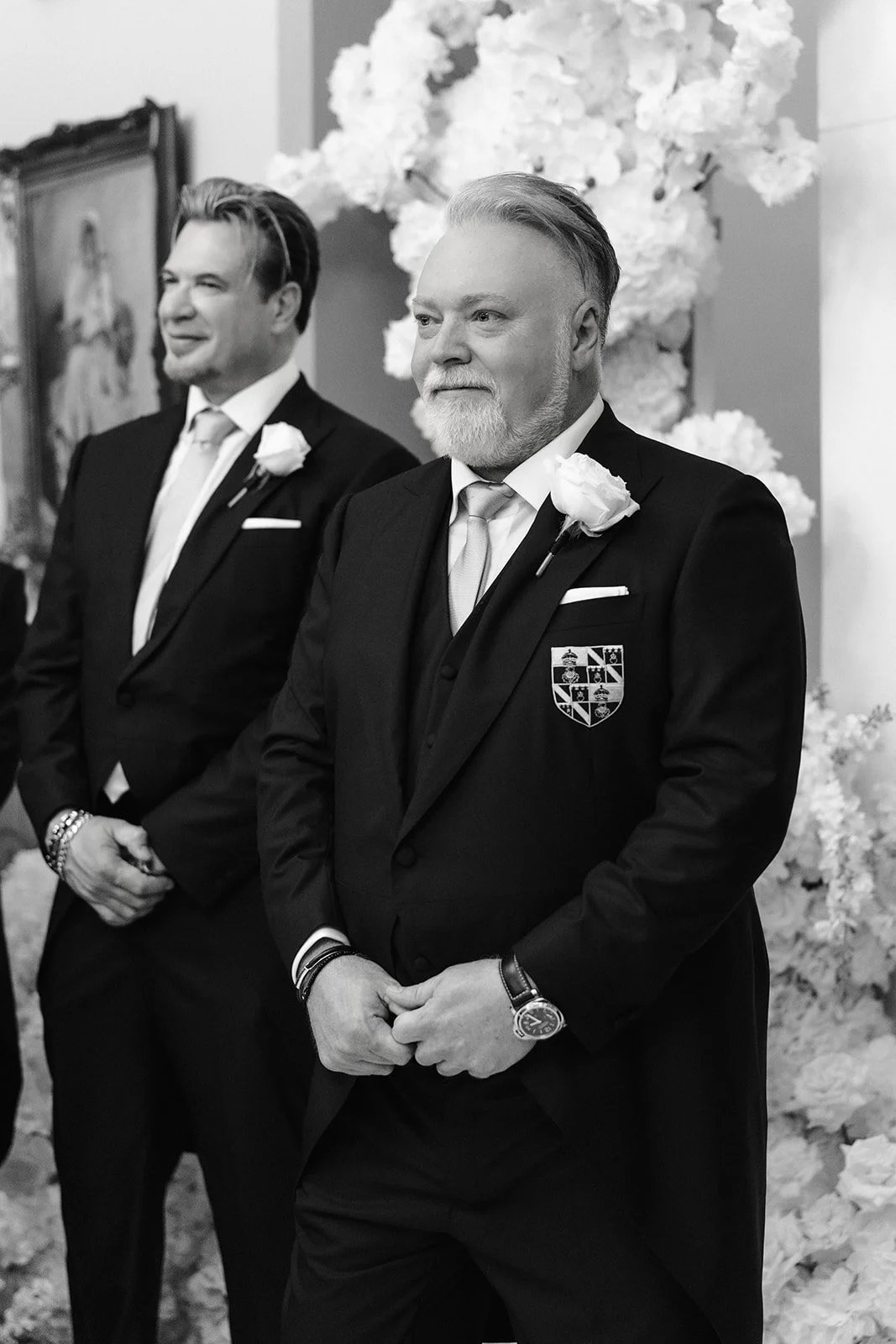 Two men in tuxedos with white roses on their lapels standing at a wedding ceremony, in front of a floral backdrop.