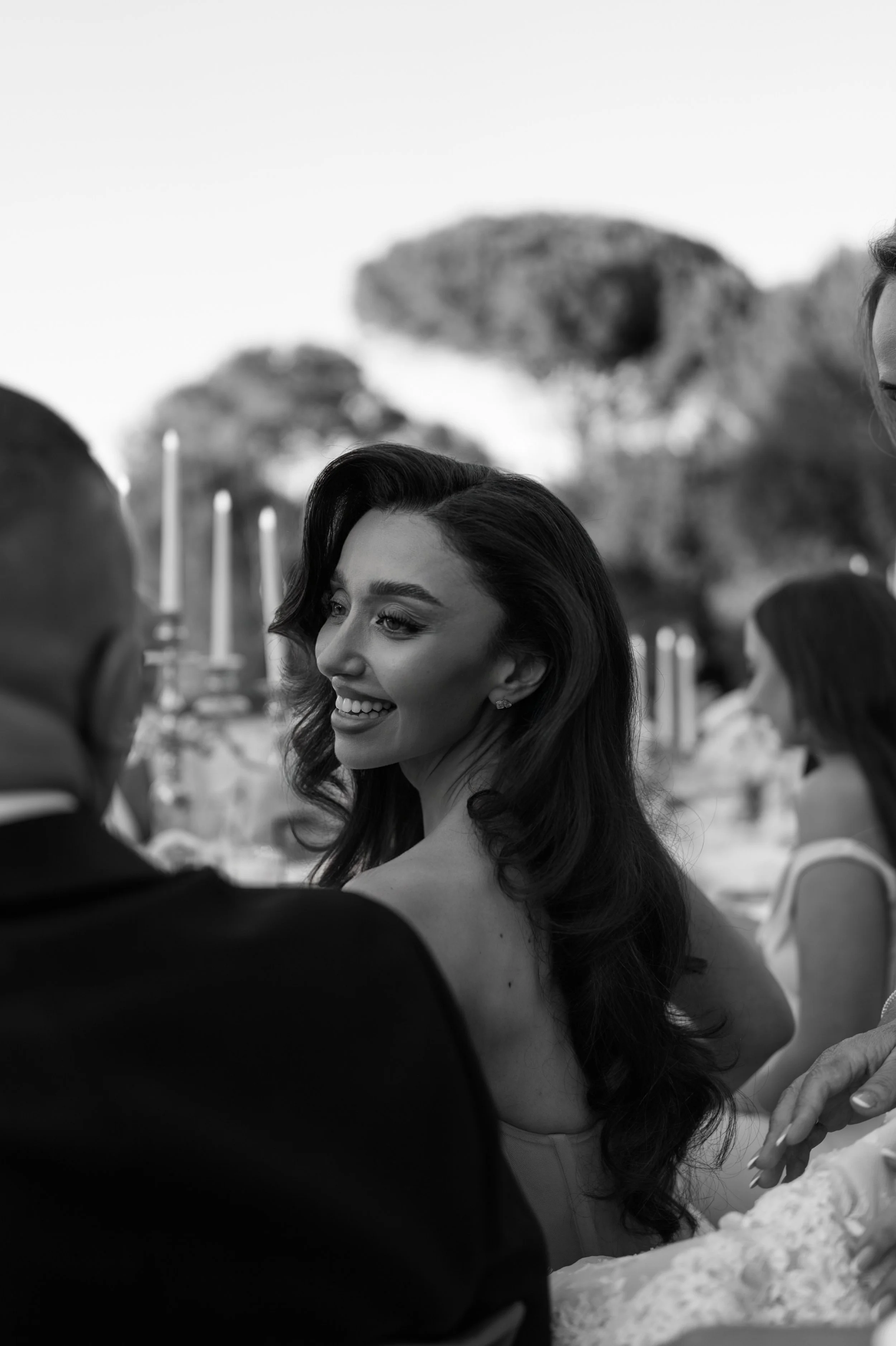 A black and white photo of a smiling woman with wavy hair attending a social event. She is wearing earrings and a sleeveless dress. There are other people around her, and a mountain is visible in the background.