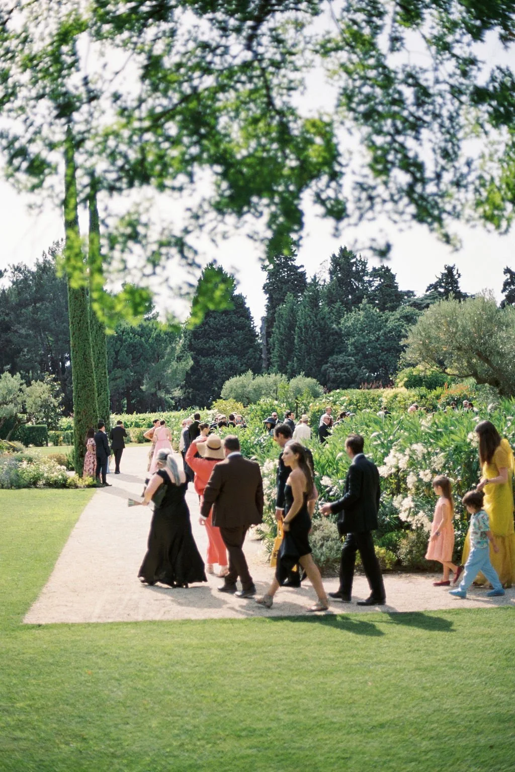 People dressed in formal attire walk along a garden path during a wedding or special event, surrounded by lush greenery and blooming flowers.