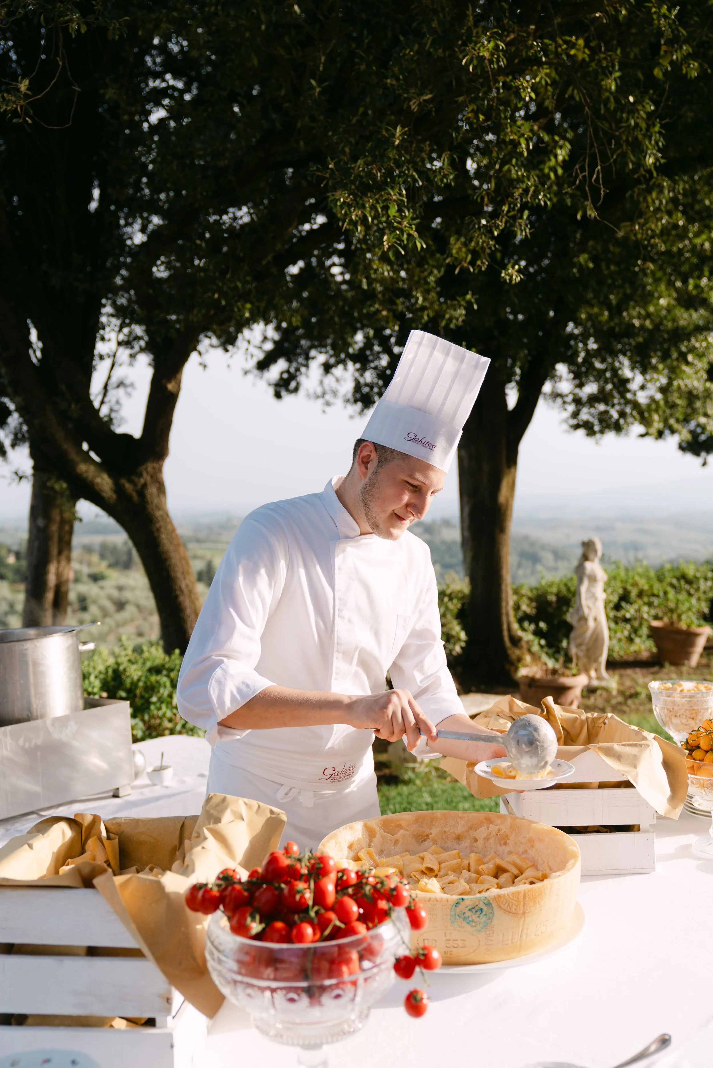 A chef wearing a white uniform and tall chef's hat is serving food at an outdoor event with trees and a scenic landscape in the background. Various food items and a bowl of cherry tomatoes are on the table.