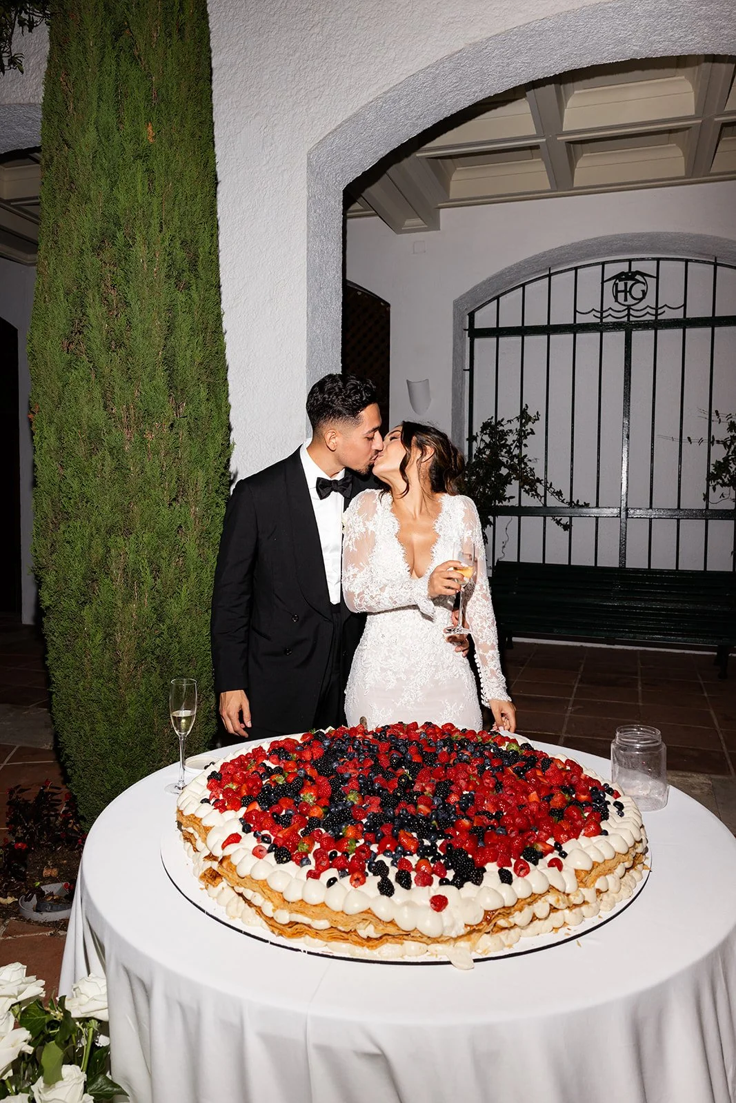A newly married couple in wedding attire sharing a kiss at the wedding reception. The bride is in a white lace gown holding a glass of champagne, and the groom is in a black tuxedo. They are standing behind a large, round layered wedding cake decorat