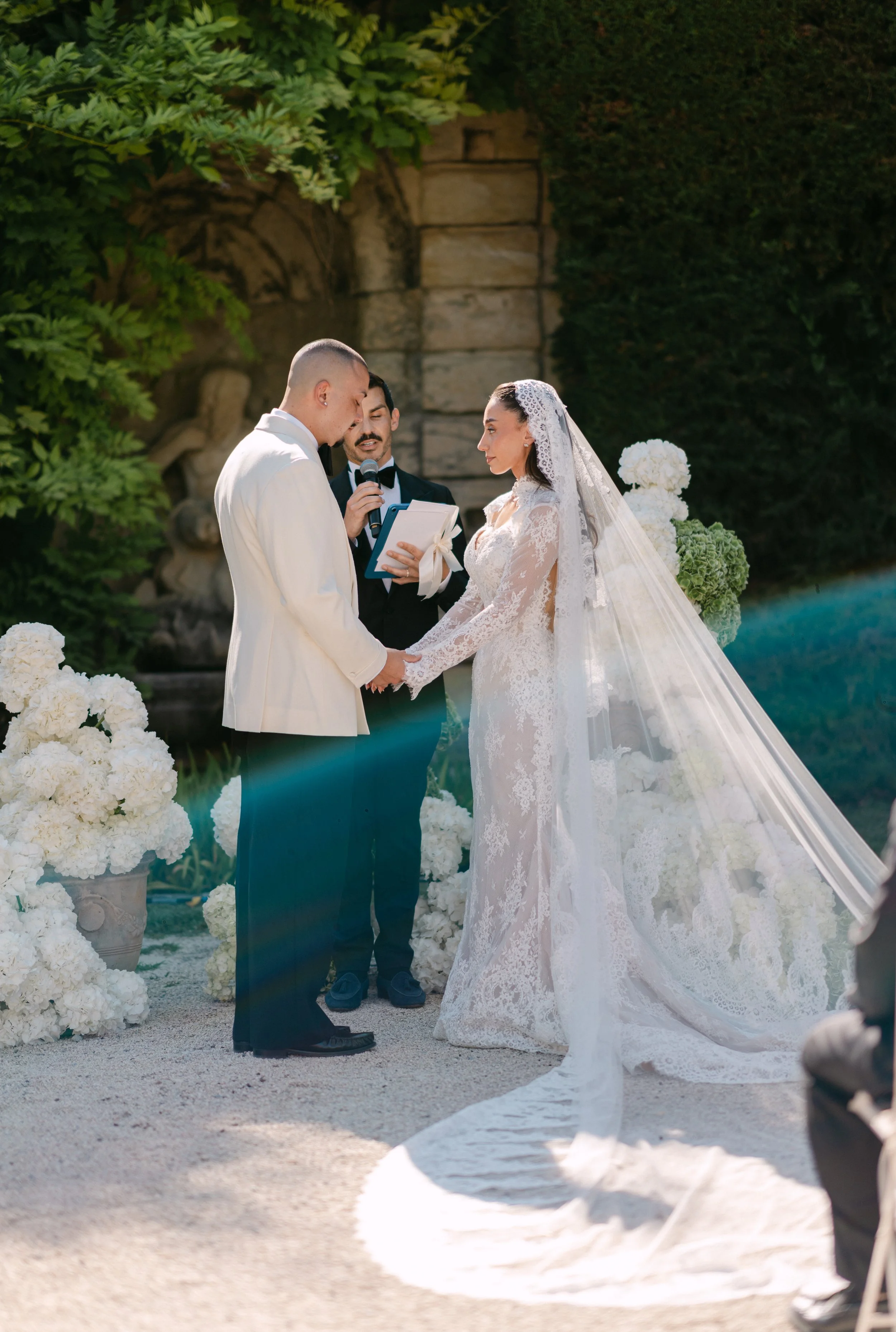 A wedding ceremony outdoors with a bride and groom holding hands, facing each other, while an officiant reads from a book. The bride is in a lace wedding dress with a long veil, and the groom is in a white tuxedo. The setting includes lush greenery a