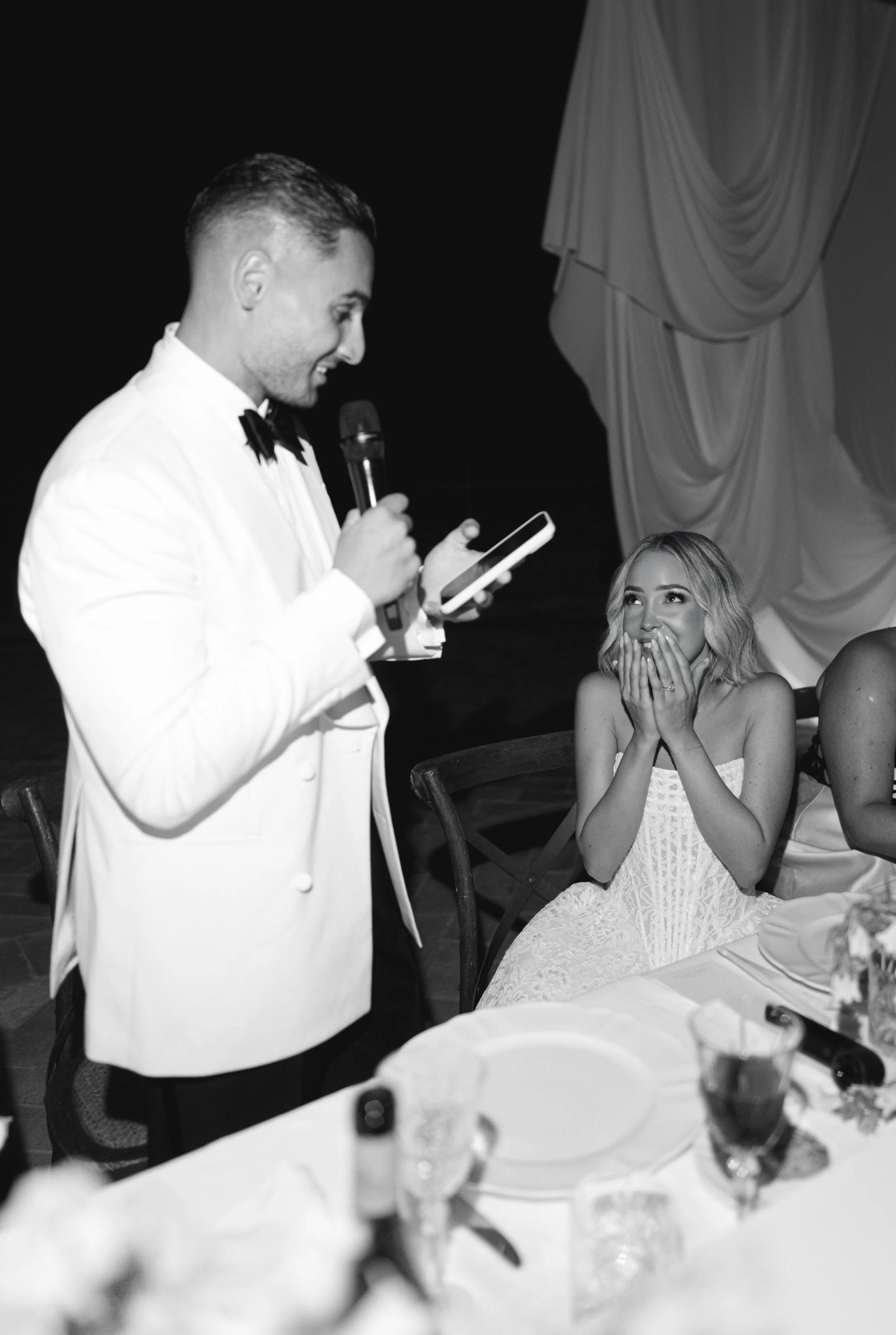 A man in a tuxedo giving a speech at a wedding reception, holding a microphone and looking at his phone, while a woman in a lace dress looks up at him with admiration and covers her mouth with her hands.