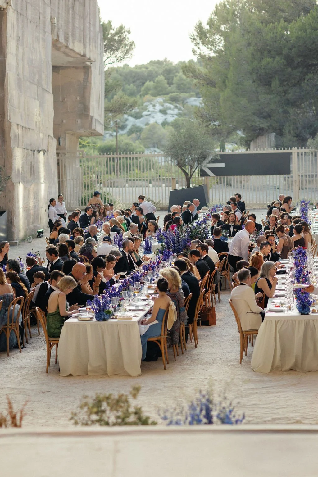 Outdoor wedding reception with long tables decorated with purple flowers and surrounded by guests in formal attire.
