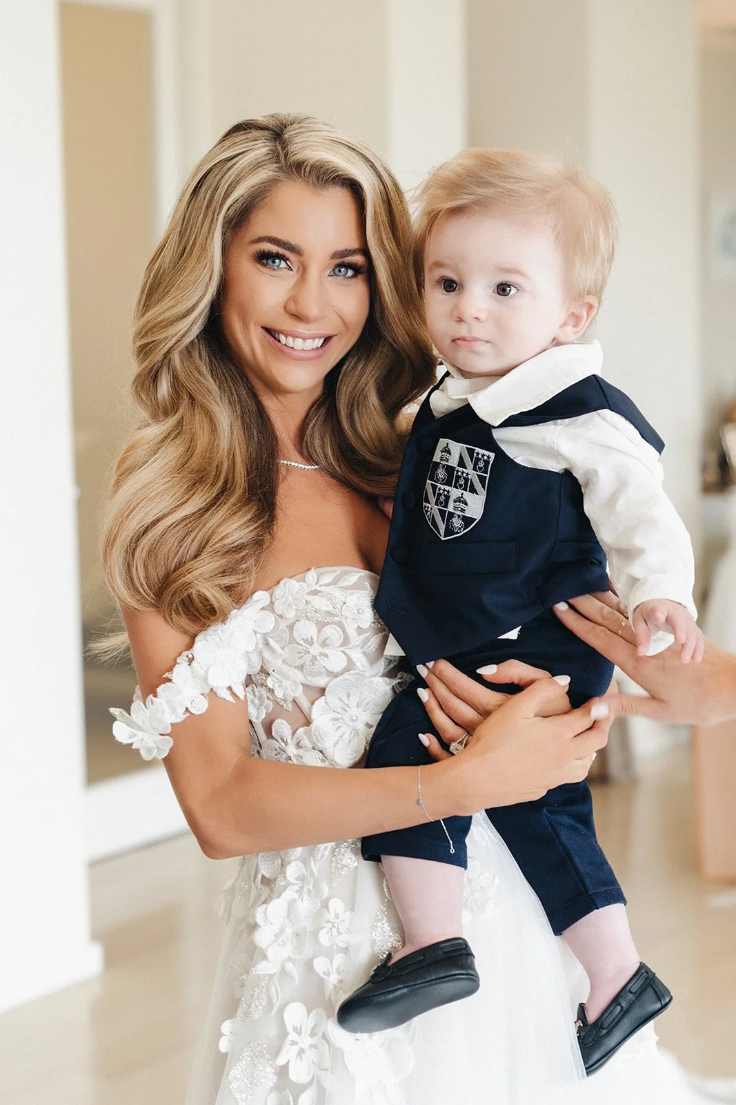 A woman in a white wedding dress holding a young boy dressed in a navy blue suit and white shirt, smiling at the camera in an indoor setting.