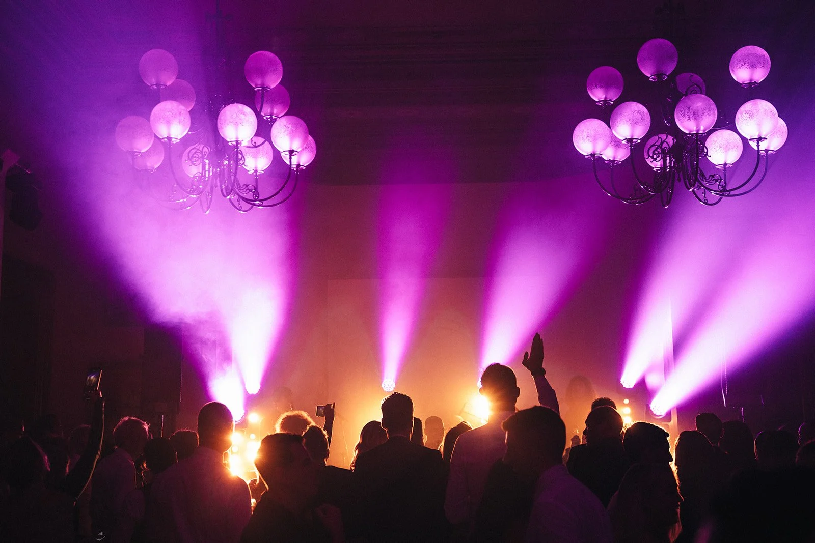 People dancing and socializing at a party or nightclub with purple stage lighting and chandeliers.