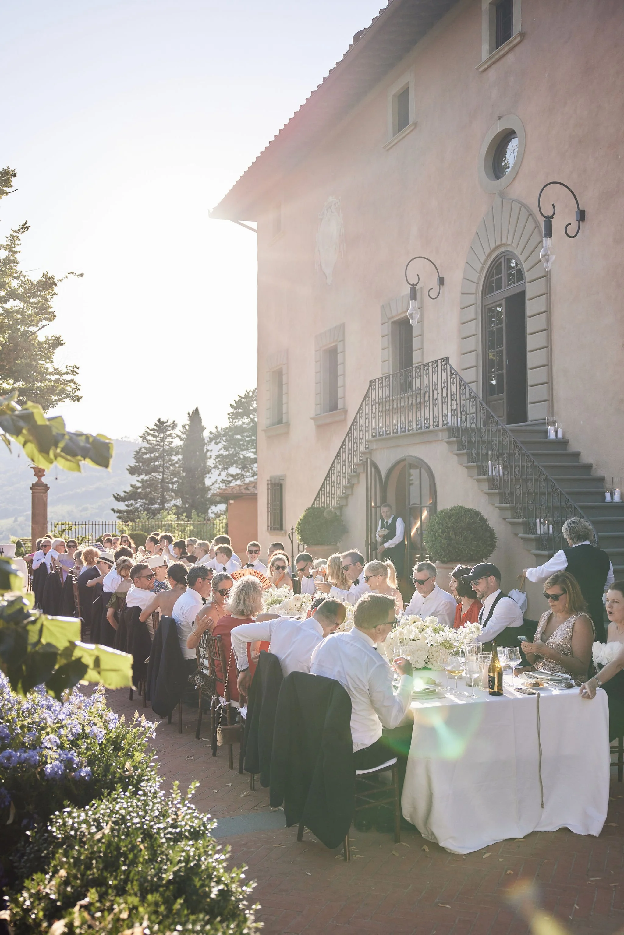 Guests gathering outside a pink stucco building for an outdoor wedding reception at sunset.