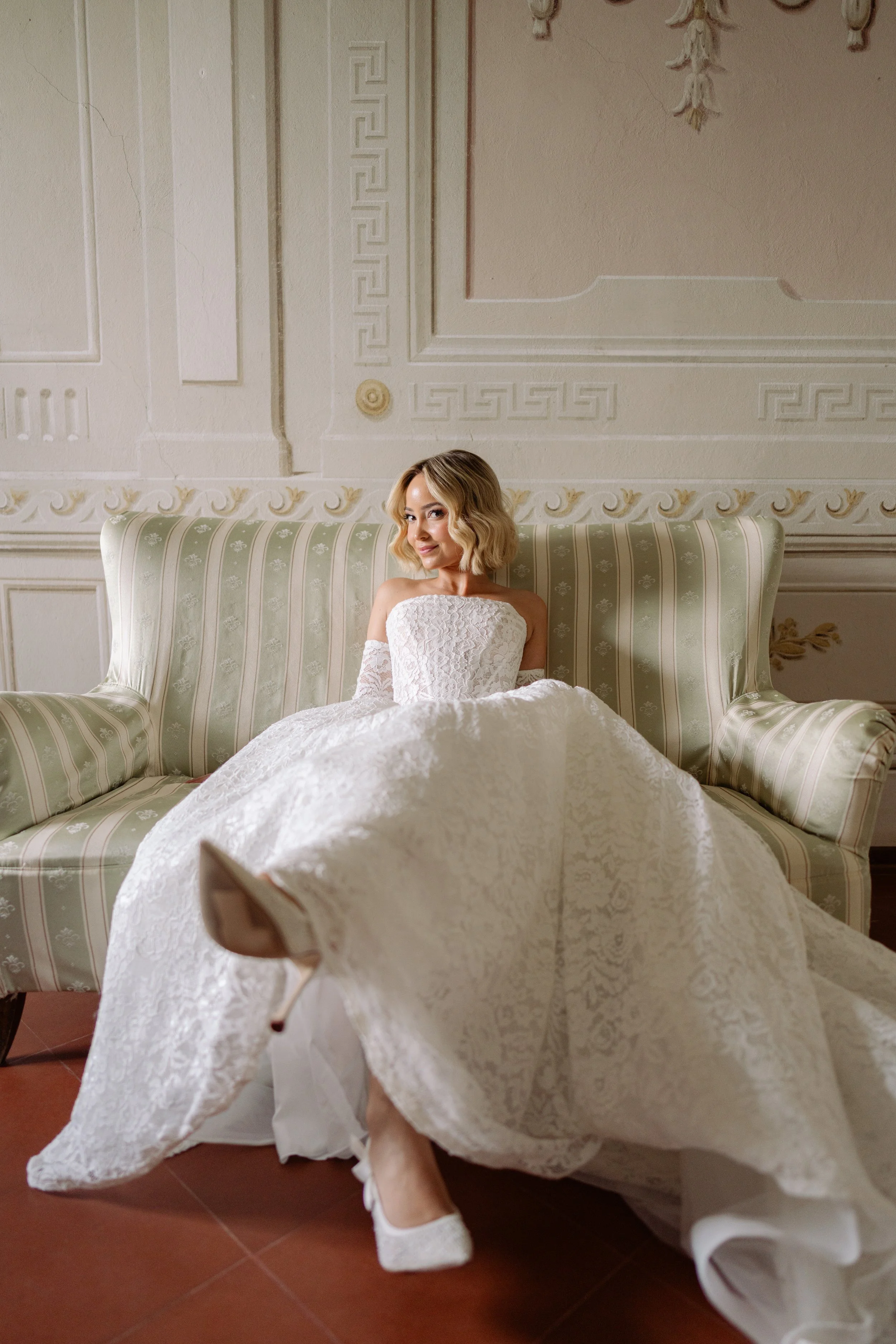 A woman dressed in a white wedding gown sitting on a couch in an elegant room with ornate wall decor.