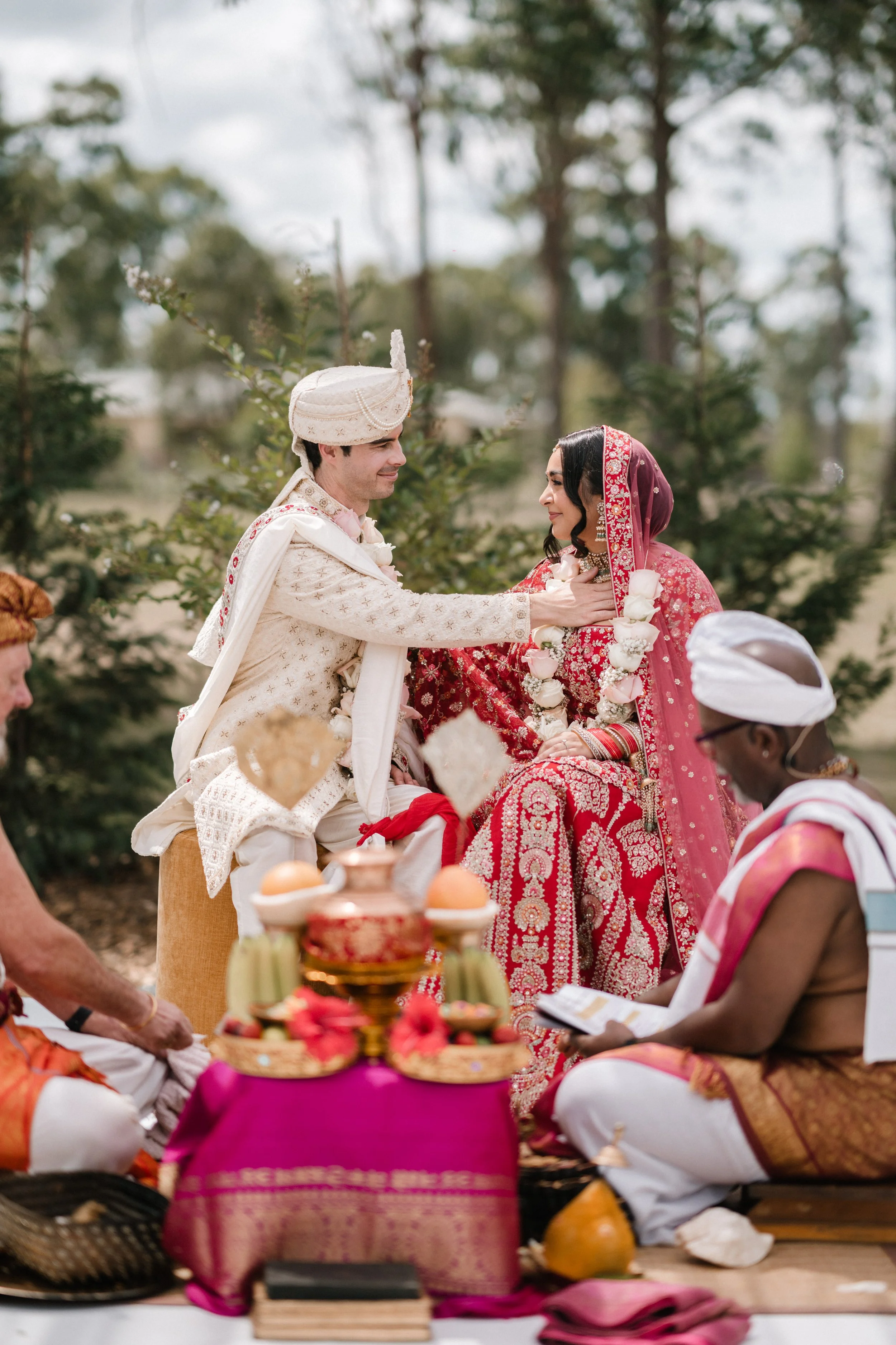 A traditional Indian wedding ceremony outdoors with a bride and groom dressed in elaborate attire, seated facing each other, with the groom placing a hand on the bride's shoulder. Several ceremonial items and participants are present, surrounded by greenery.