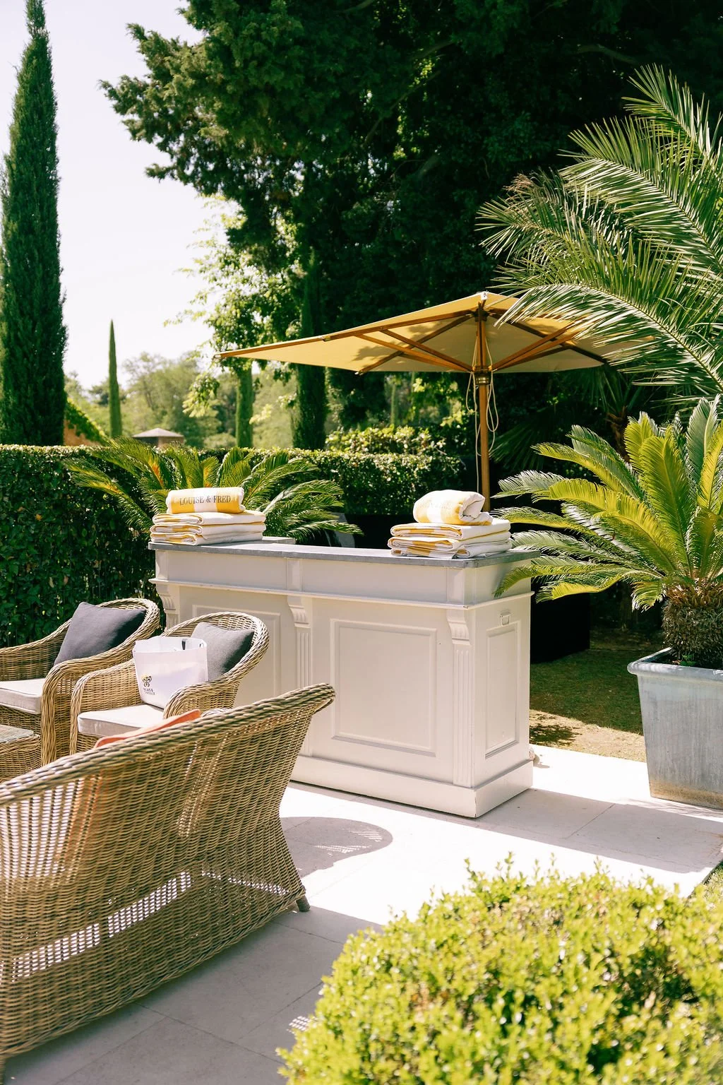 An outdoor patio with wicker chairs, a white bar counter, and a large yellow umbrella, surrounded by lush green plants and trees.