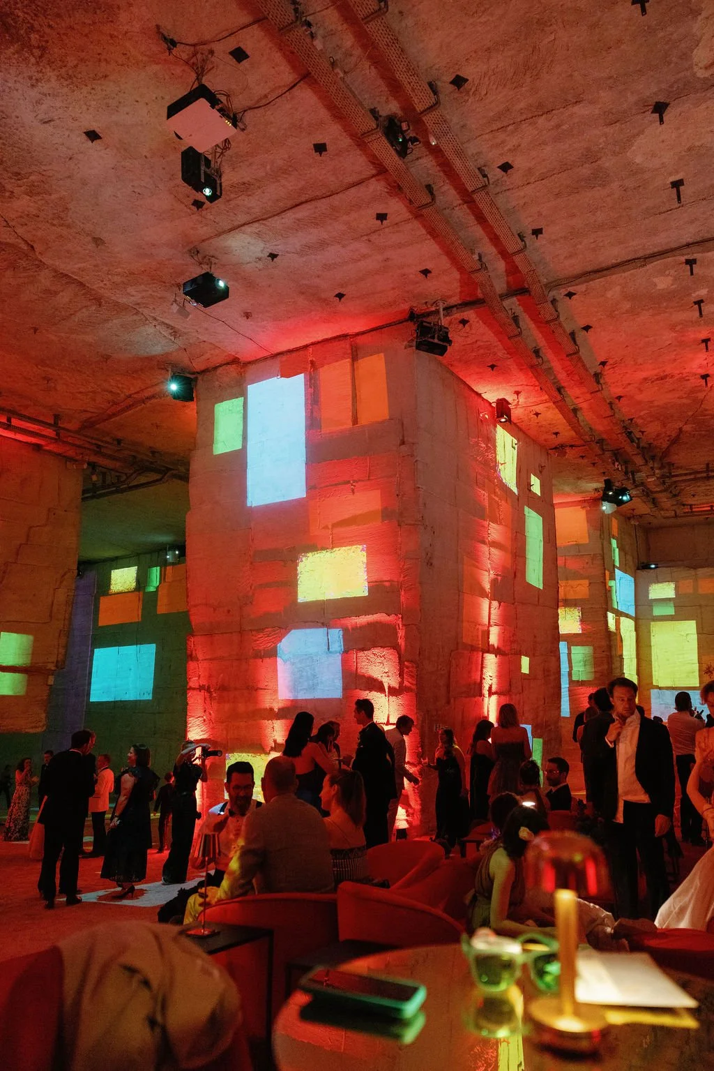 A lively indoor party scene with people dancing and socializing under colorful projected lights on stone walls, with some seated around tables.