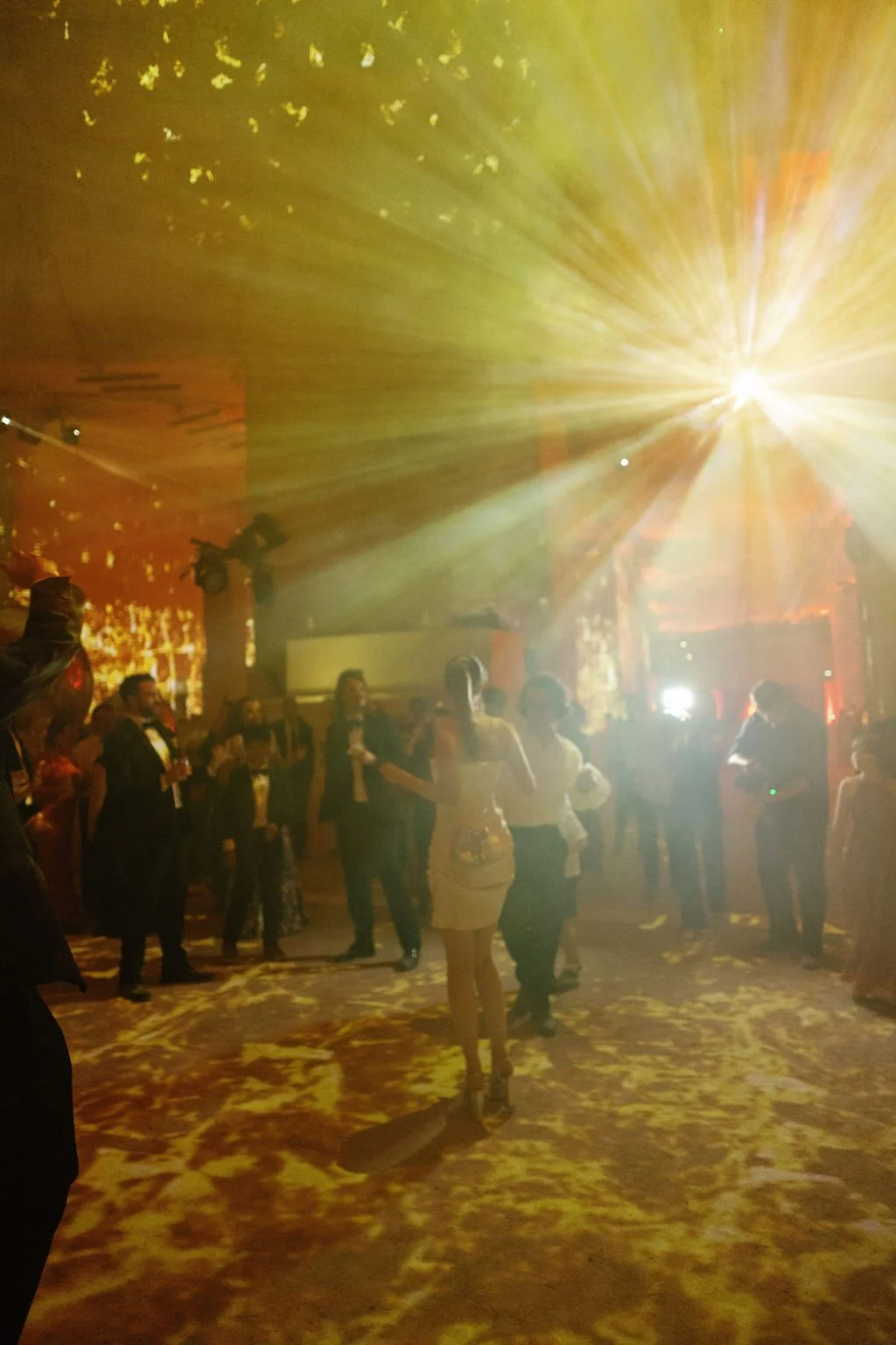 People dancing and socializing at a nightclub or party with bright yellow stage lighting and a patterned dance floor.