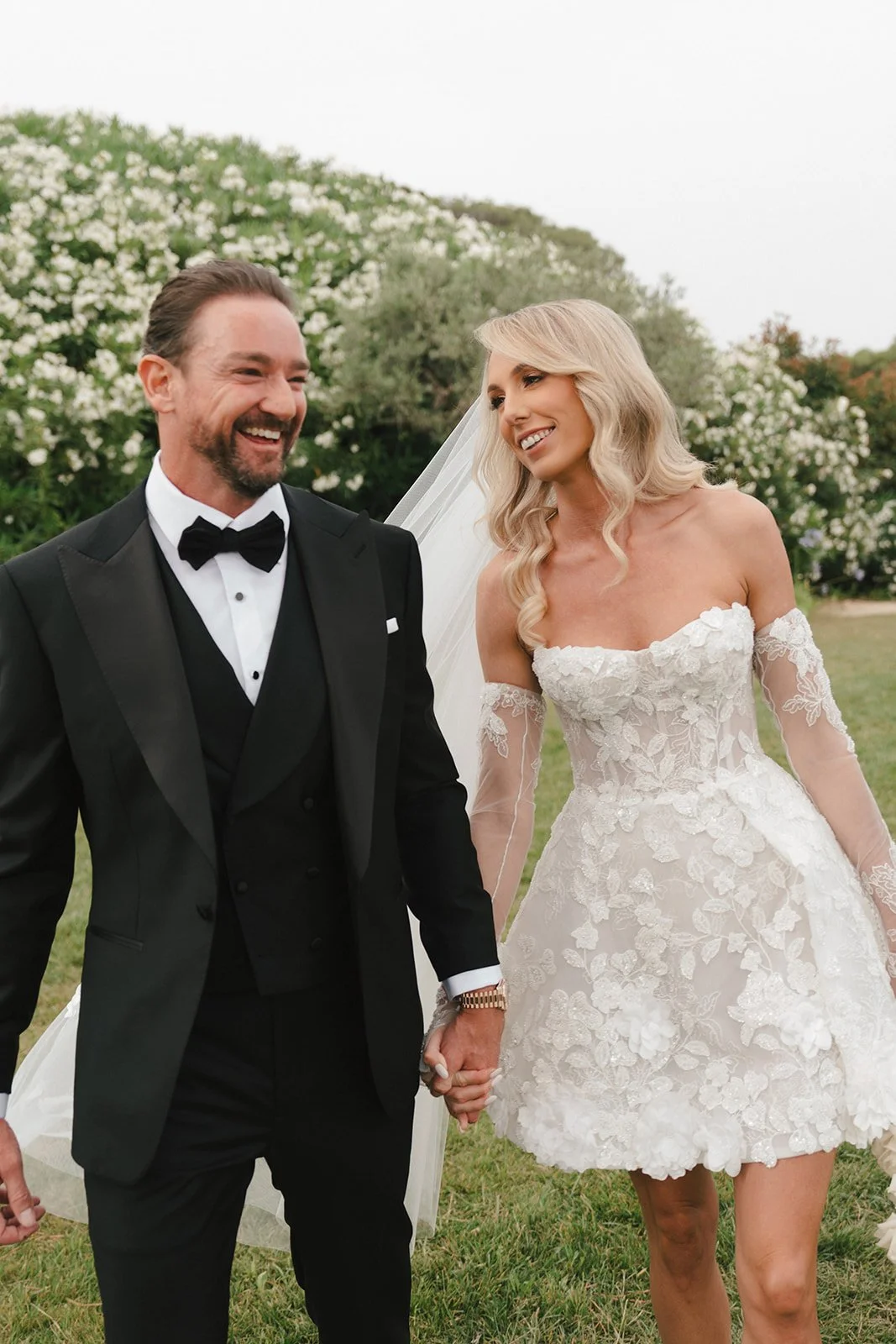 A newly married couple holding hands and smiling outdoors, with the woman in a white, lace off-shoulder wedding dress and the man in a black tuxedo, surrounded by greenery and blooming trees.