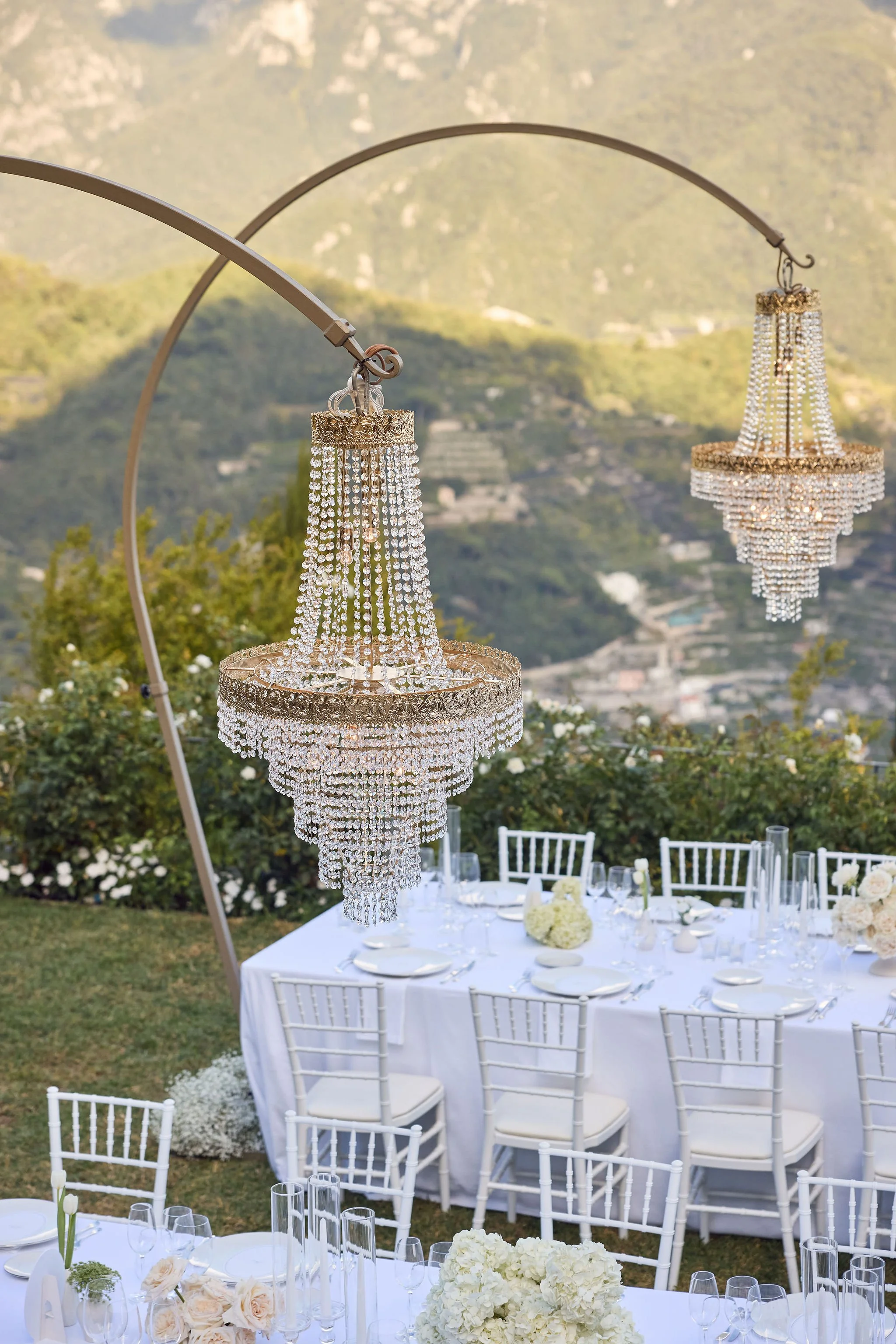 Chandeliers hanging above a decorated outdoor dining table with white chairs and floral centerpieces, set against a scenic mountain backdrop.