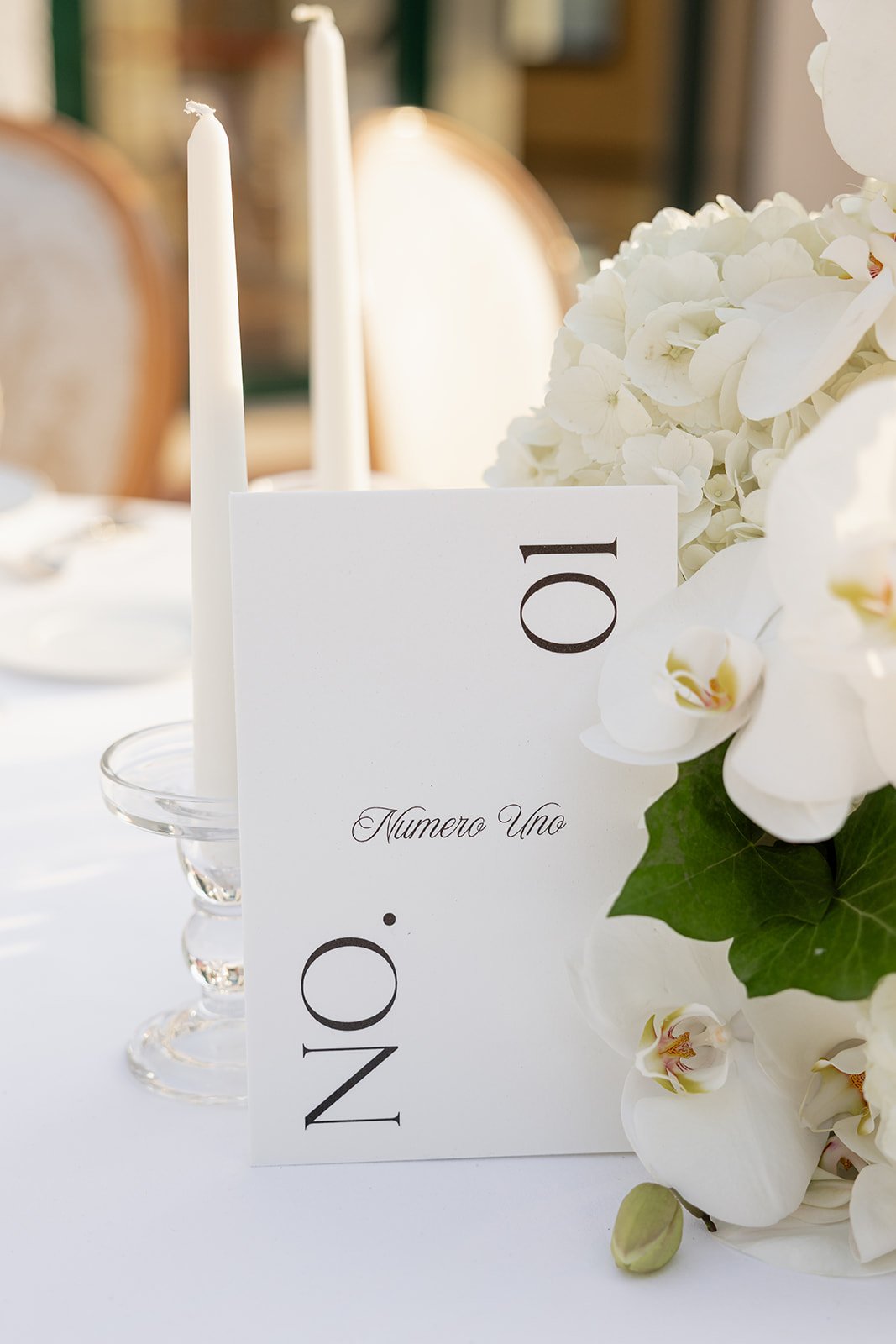 Elegant table setting with a white numbered card reading '10', white candles, and a floral arrangement of white orchids and hydrangeas at a formal event or wedding.