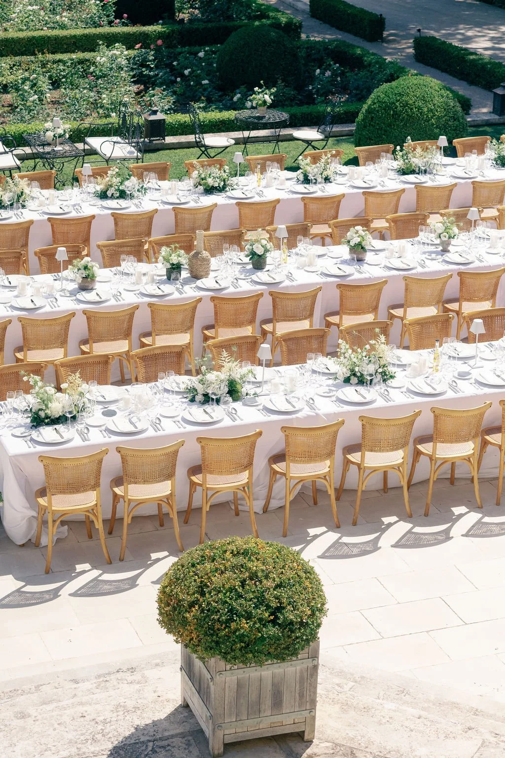 Elegant outdoor dining setup with long tables covered in white tablecloths, decorated with white floral centerpieces, and surrounded by wicker chairs in a garden setting.