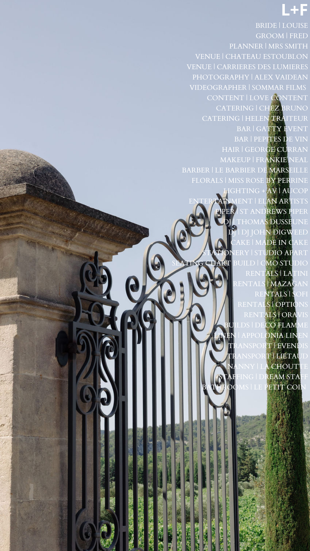 A decorative black wrought iron gate attached to a stone pillar, with a scenic landscape of trees and hills in the background.
