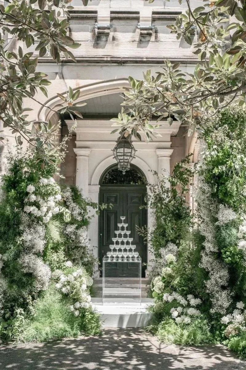 Champagne tower in front of a black doorway framed by greenery and white flowers.
