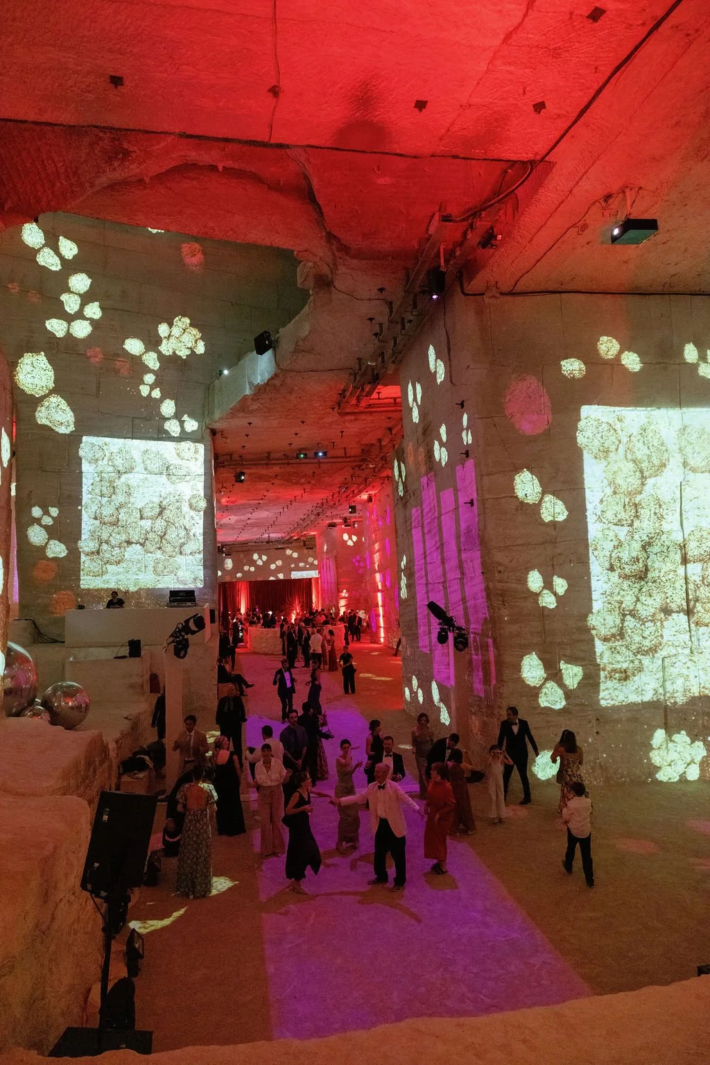 People dancing and socializing in a large, industrial-style event space with high ceilings, projected abstract visuals on the walls, and colorful lighting.