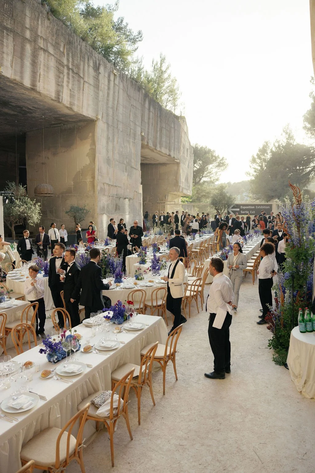 An outdoor formal event with guest dressed in tuxedos and suits, seated at elegant tables decorated with purple flowers, set under a large concrete structure with trees and a bright sky in the background.