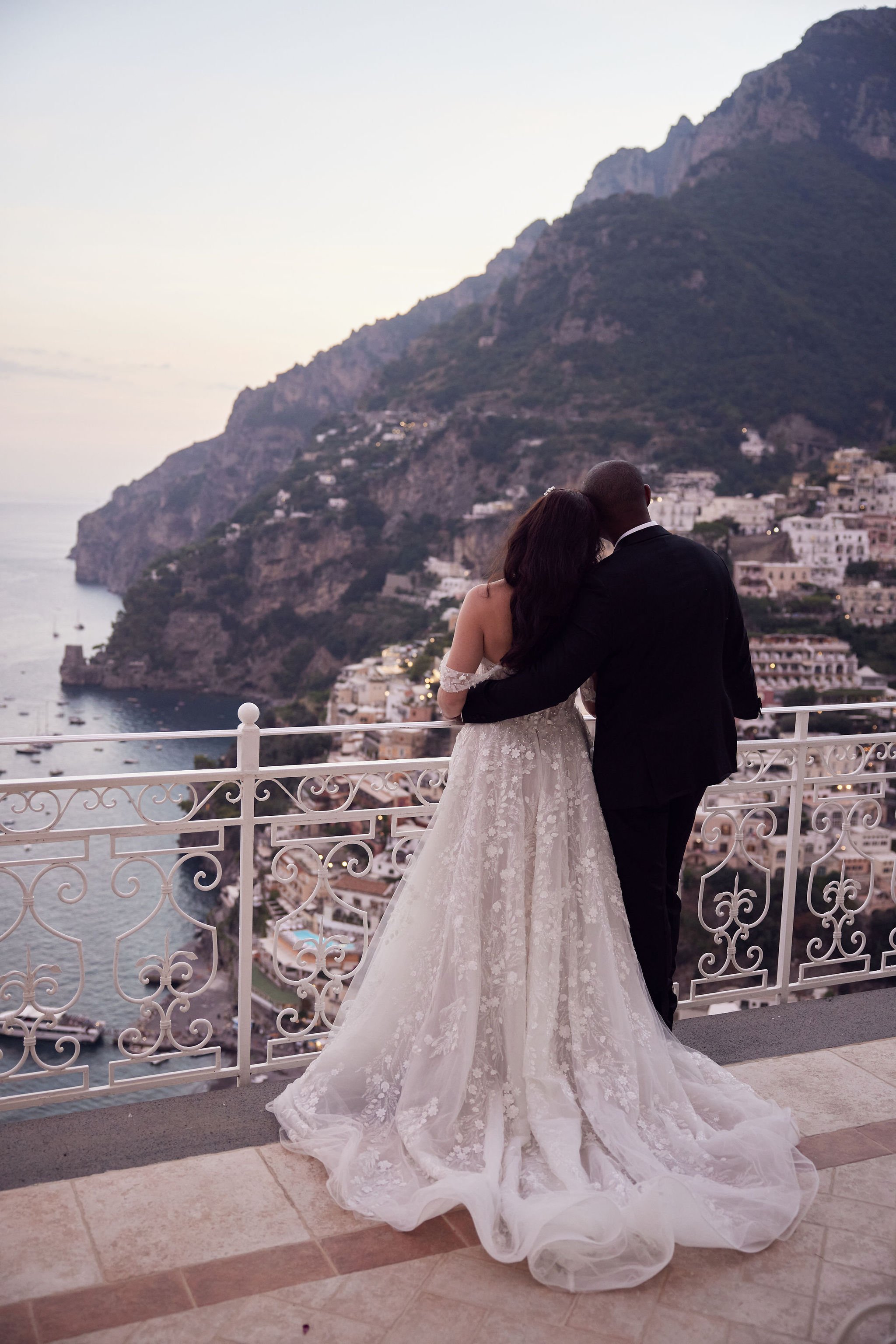 Couple in wedding attire standing on a balcony overlooking a coastal town with mountains in the background at sunset.