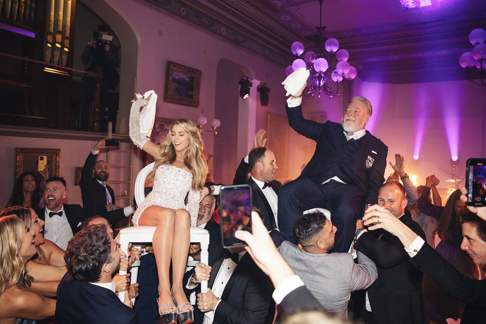 People celebrating at a wedding reception, with a woman in a white dress sitting on a chair and a man in a tuxedo sitting on a chair lifted by others, waving white napkins, amid a lively crowd with purple lighting and chandeliers.